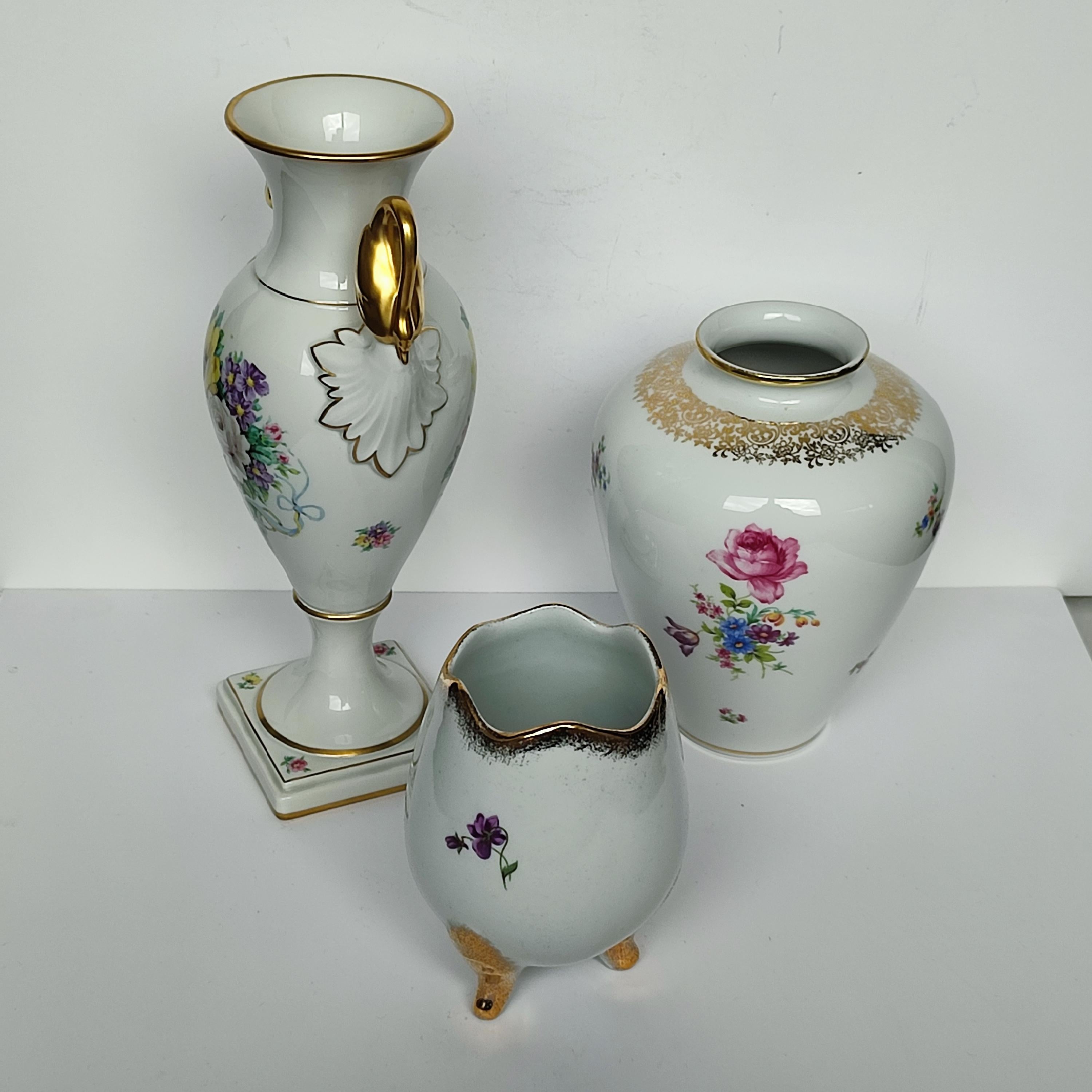 Dieses kuratierte Set aus drei exquisiten deutschen Porzellanvasen aus der Mitte des Jahrhunderts ist eine raffinierte Symphonie aus Form, Kunstfertigkeit und Eleganz, die Ihren Raum aufwertet. Jedes Stück ist mit lebhaften Blumenmotiven handbemalt