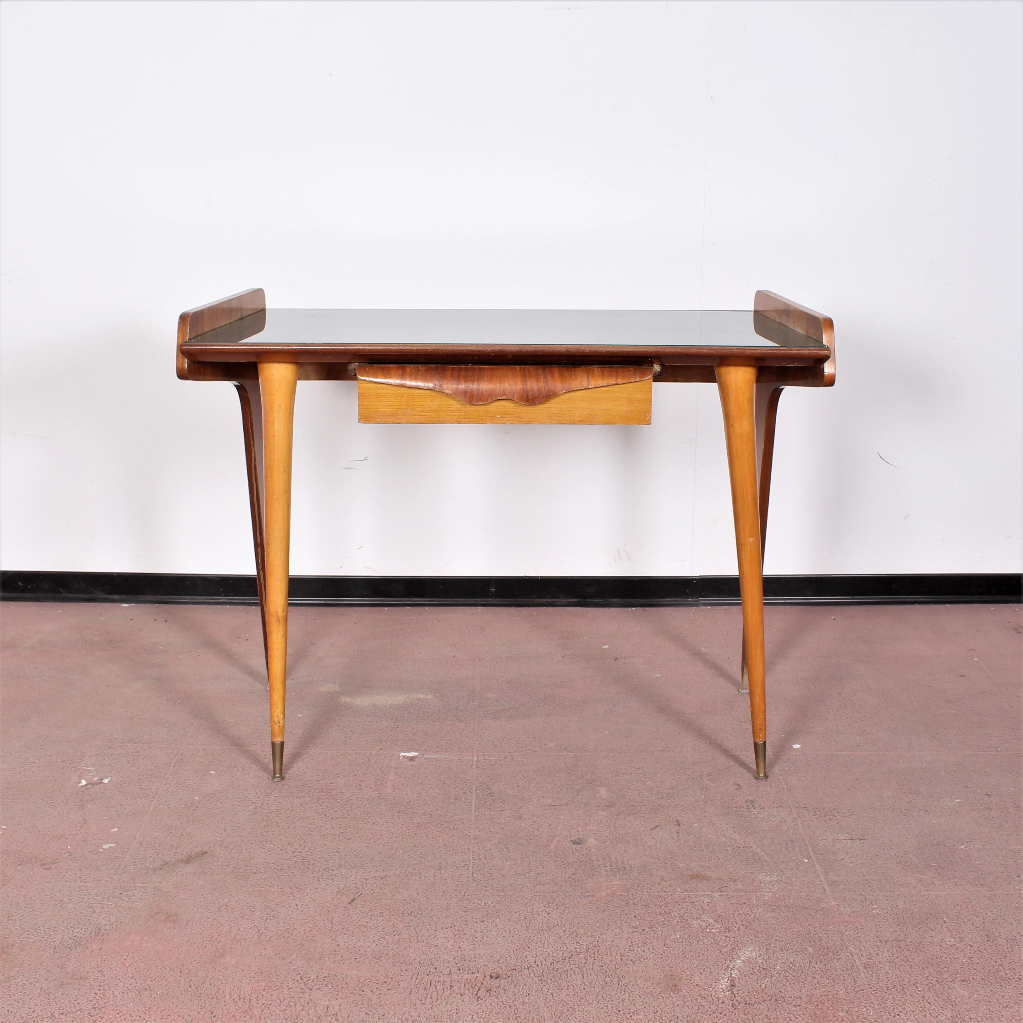 Very original and beautiful maple wood console table with front drawer, brass frontal feet and green colored glass top. Striking asymmetry in the layout and shape between the front legs, conical and closer together, and the rear ones, wider and