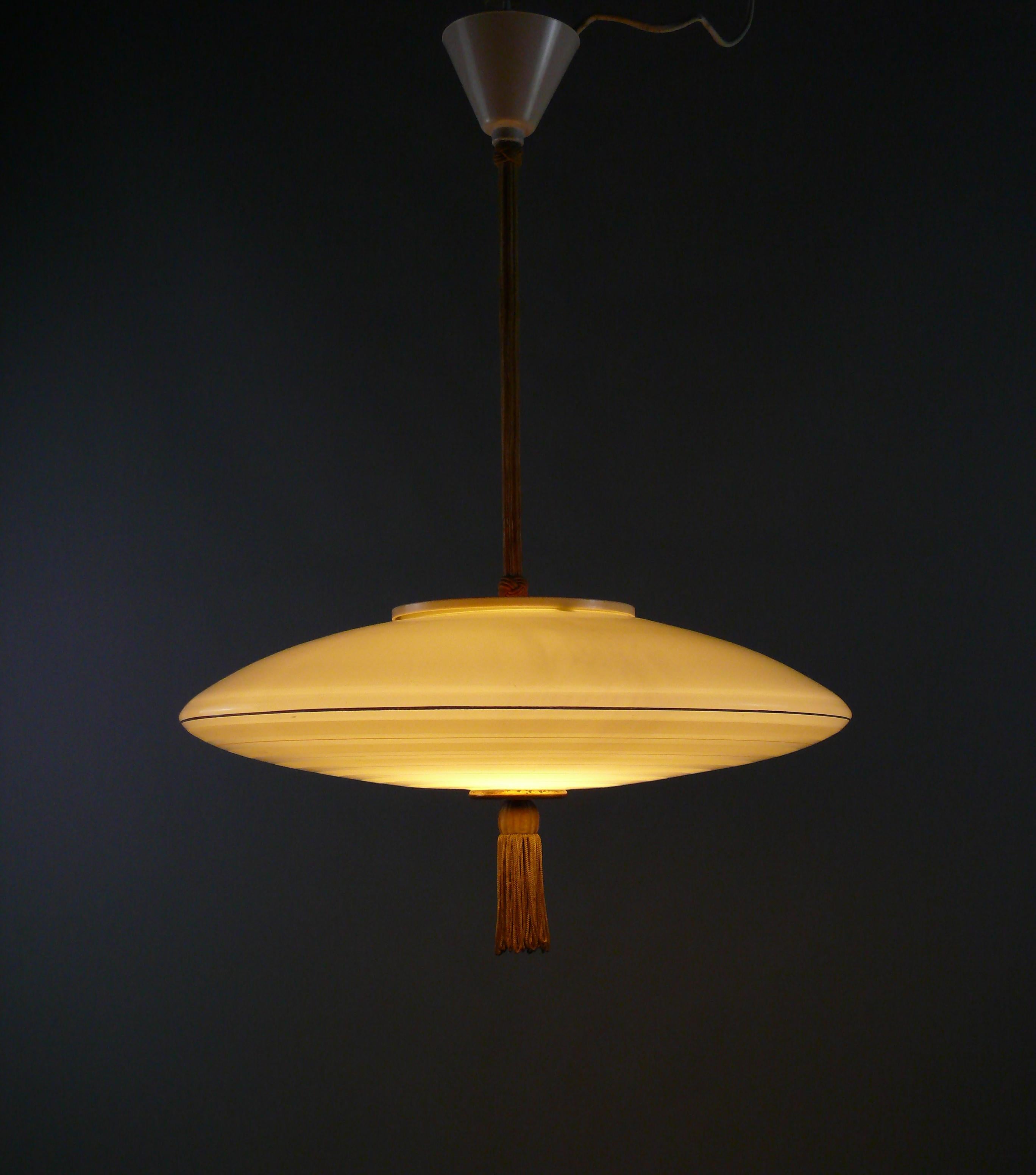 Large rod pendant light from the 1950s with a beige glass shade, coated rod and plastic suspension.

The ufo-shaped glass shade has radial-shaped, gold-colored decorative strips and frosted areas. The lower end button is made of brass and has a