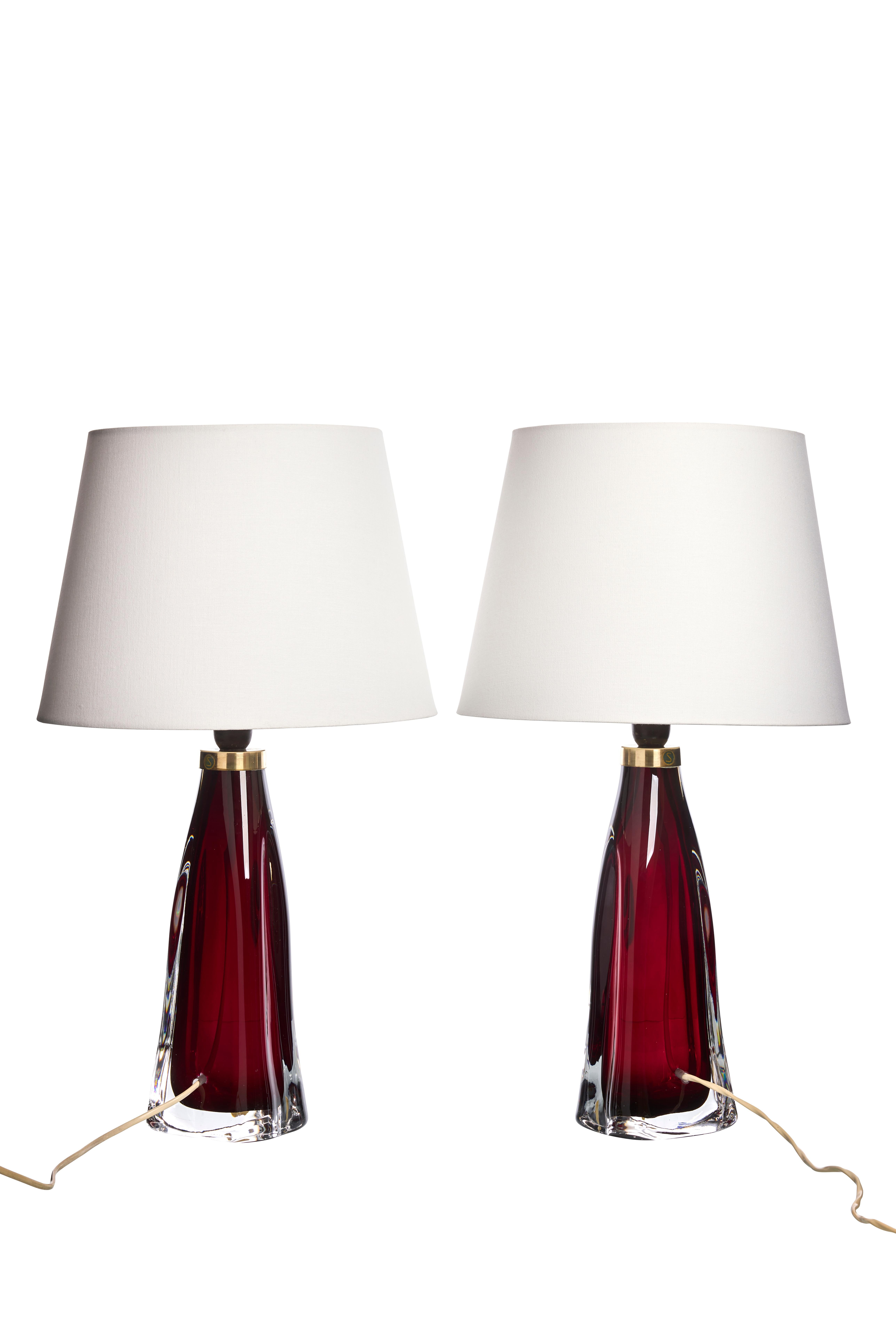 These table lamps are a pair of Mid-Century pieces designed by Carl Fagerlund for Orrefors in Sweden during the 1960s. Each lamp features a striking red glass base with a clear glass bottom, giving them a sleek and elegant appearance. The bases are