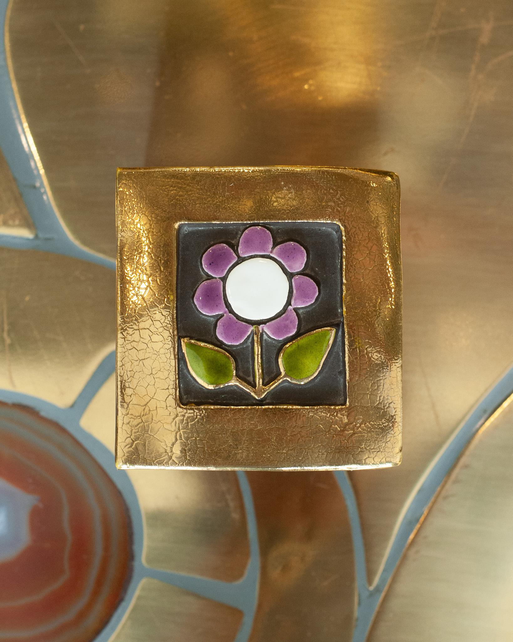 Un'eccezionale scatola in ceramica smaltata di metà secolo con bordo in oro liquido e fiore viola, opera della famosa artista Mithé Espelt. Il coperchio della scatola è in ceramica con una base in legno annerito.
Nata nel 1923, Mithé Espelt era