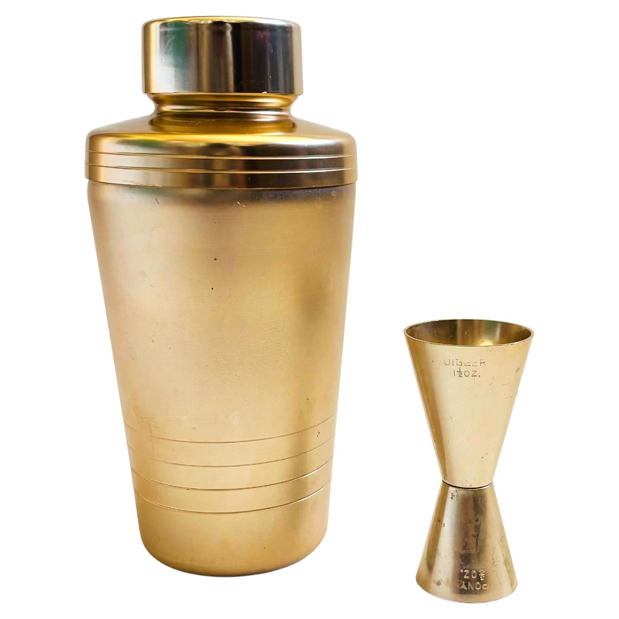 A Mid Century gold-tone aluminum two piece cocktail set including a shaker and a 2 sided jigger. The shaker is matte gold in coloring with shiny gold bands and a cap for contrast. 
Measurements:
Cocktail Shaker: 4