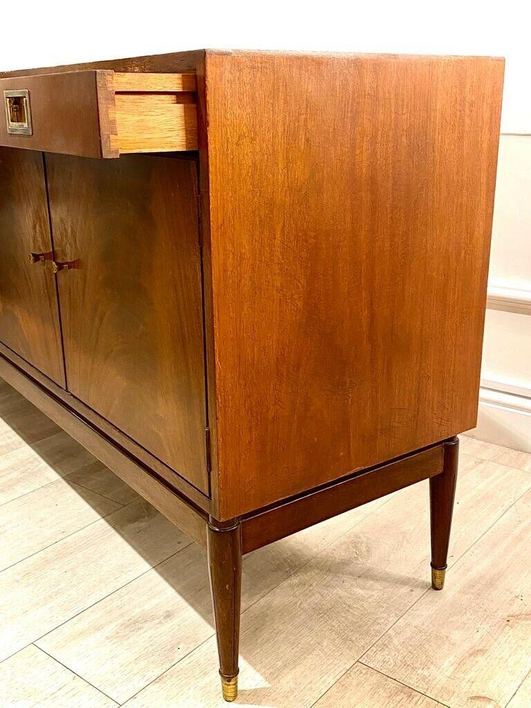 A beautiful Greaves and Thomas teak and flame mahogany sideboard with brass coated handles and feet caps. This piece consists of 4 drawers above four cupboard doors which conceal shelving.

Original paper label on the back stating this piece