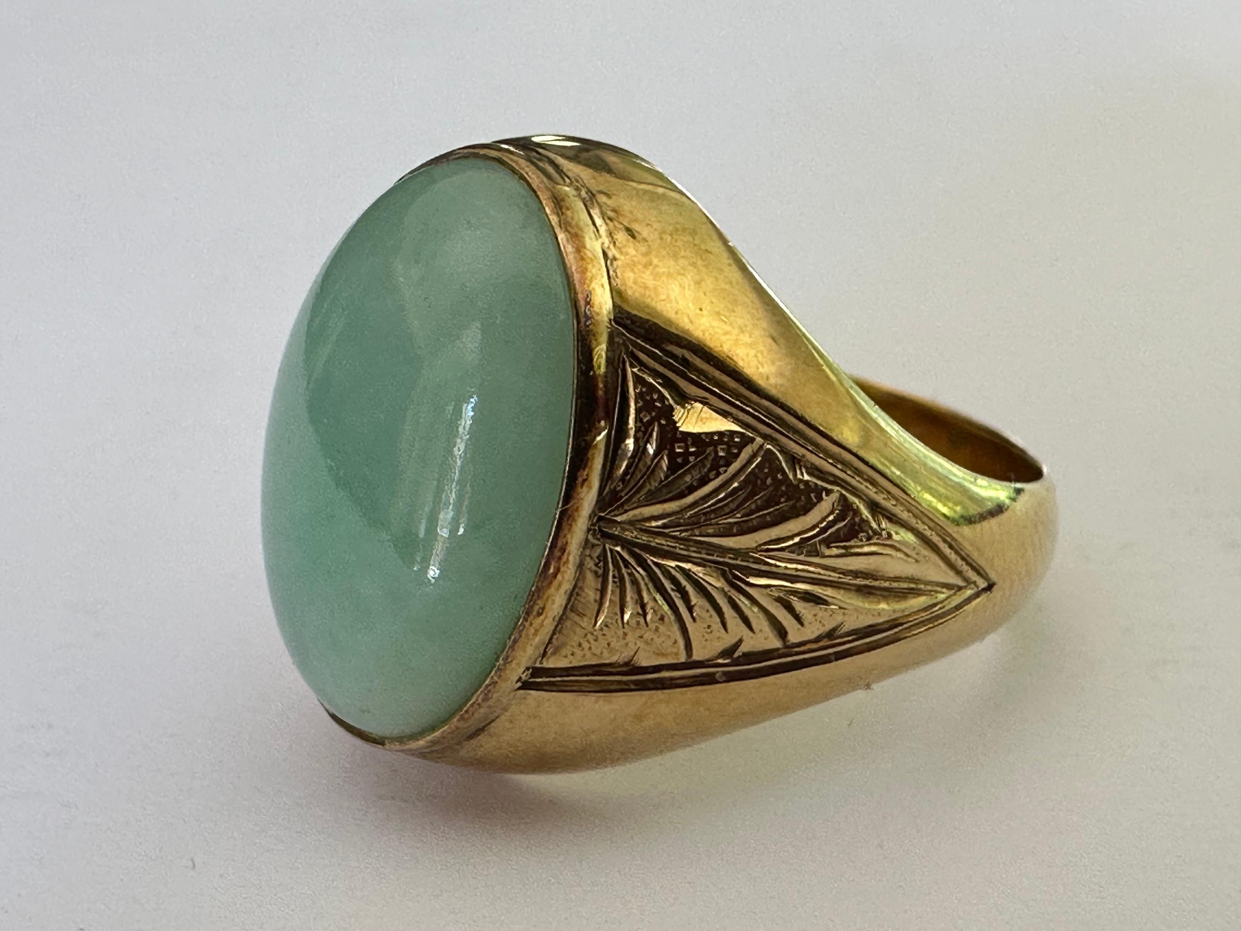 A large light green jade oval-shaped cabochon measuring approximately 17x13mm centers this mid-century ring crafted from 14K yellow gold, hand-engraved with an exquisite feather-like pattern on the shoulders. Circa 1950s. 
