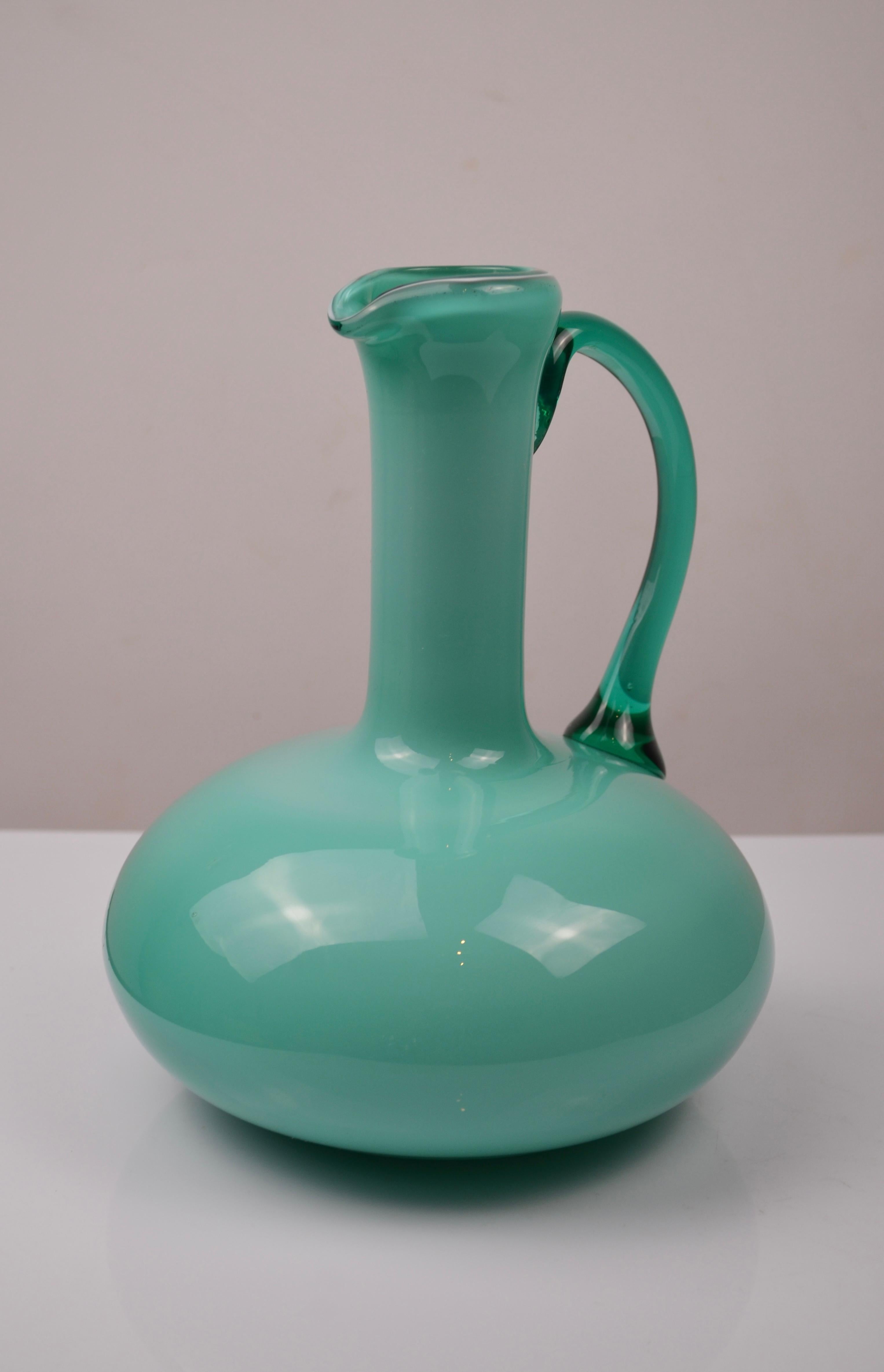 Elegante carafe vintage en verre opalin vert, soufflée à la bouche et finie à la main, avec un corps gracieusement arrondi et un bec légèrement évasé. La poignée, en verre légèrement translucide, contraste magnifiquement avec le corps opalin,