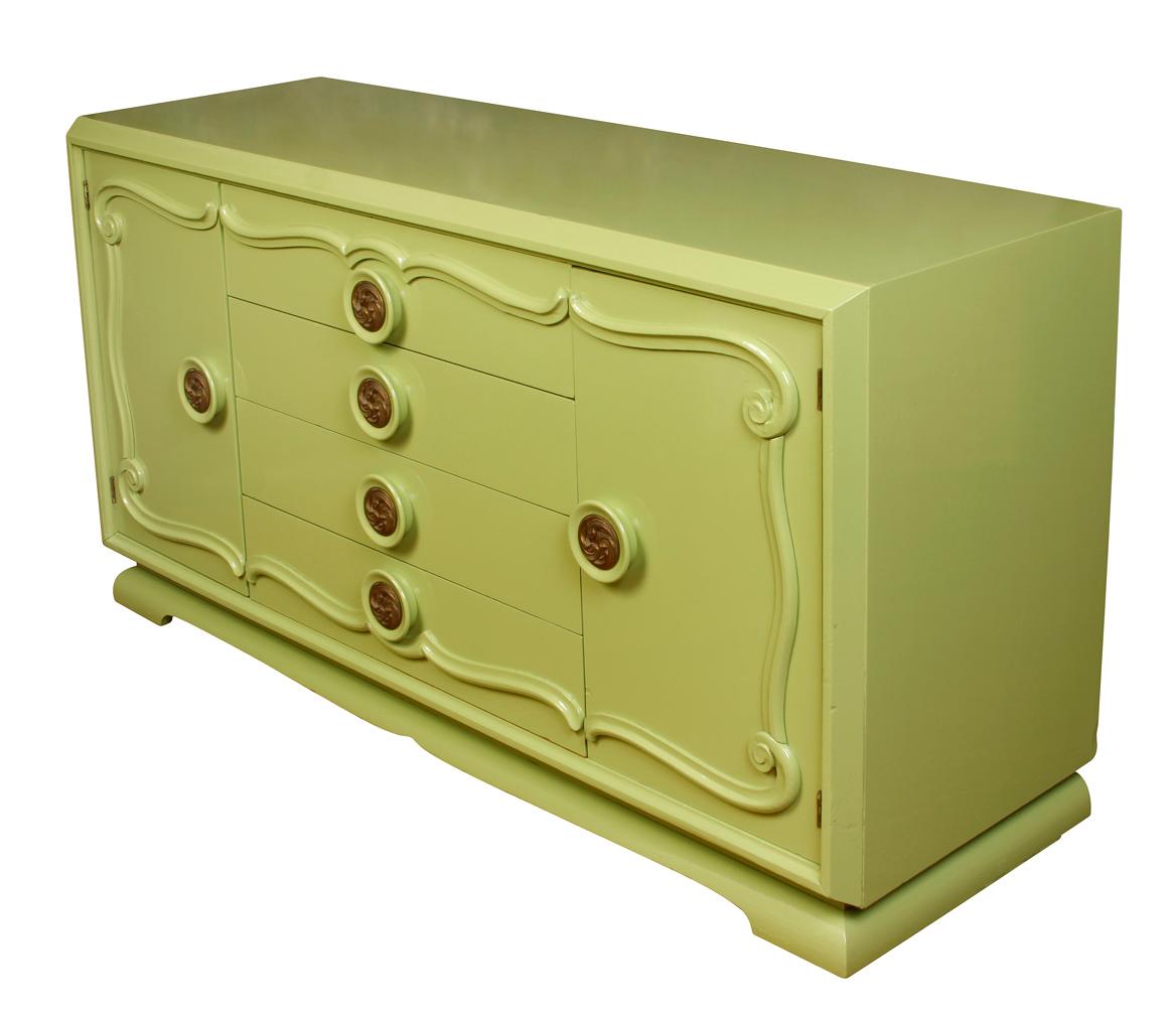 A midcentury light green painted credenza, circa 1960. The credenza has circular brass pulls with scroll detail. The front of the credenza is detailed with swirled molding. On each side is a door that opens to a drawer and two shelves. In the center