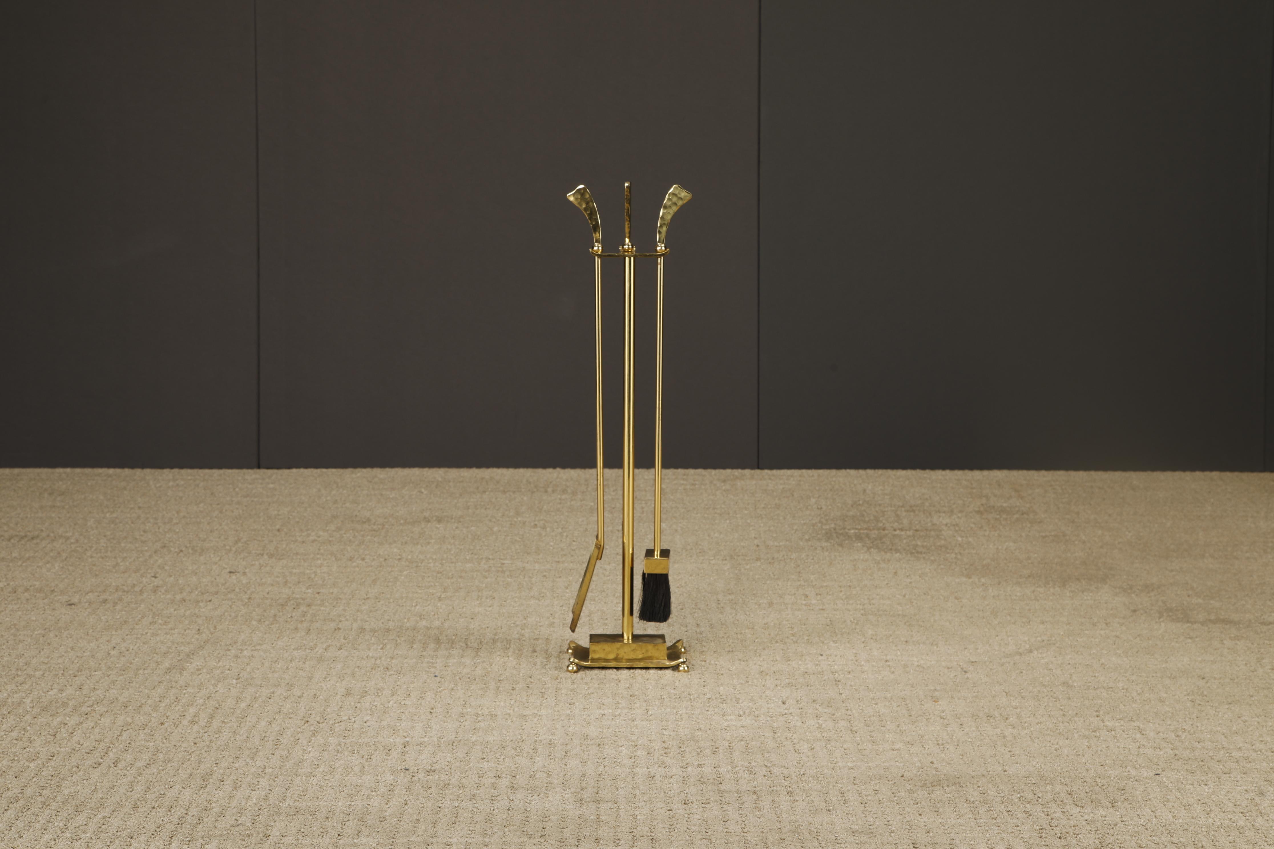 A sculptural and richly detailed mid-century modern fireplace tool set in polished hammered brass. The set includes a stand, brush, poker, and shovel, all suspended from a decorative rack with curled handles and orb finials. The hammered texture