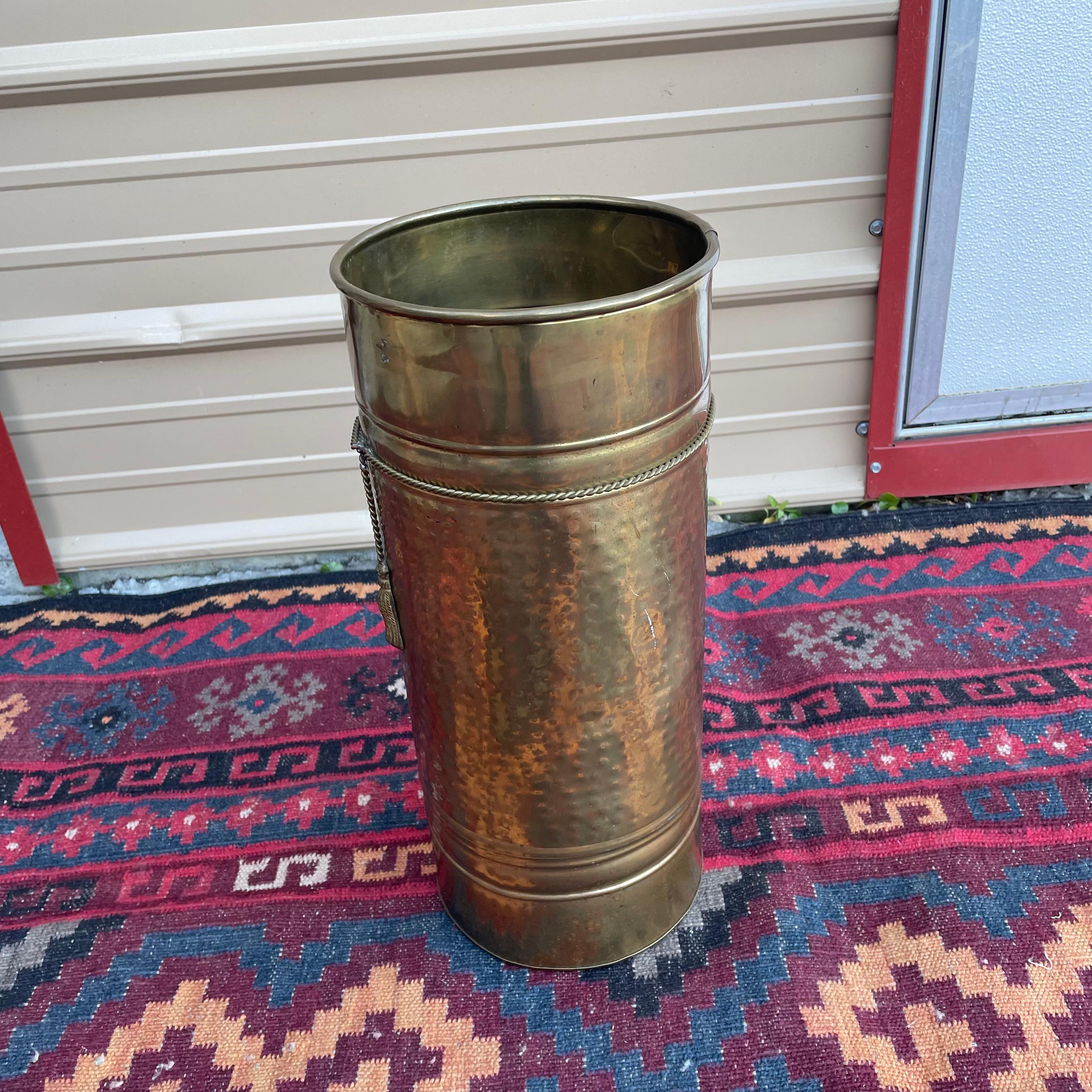 Vintage hammered brass umbrella stand with a draped brass rope design tied around it. In overall good shape. There are some dull spots in the brass finish along with some patina around the rope design. A few small dents. All wear is consistent with