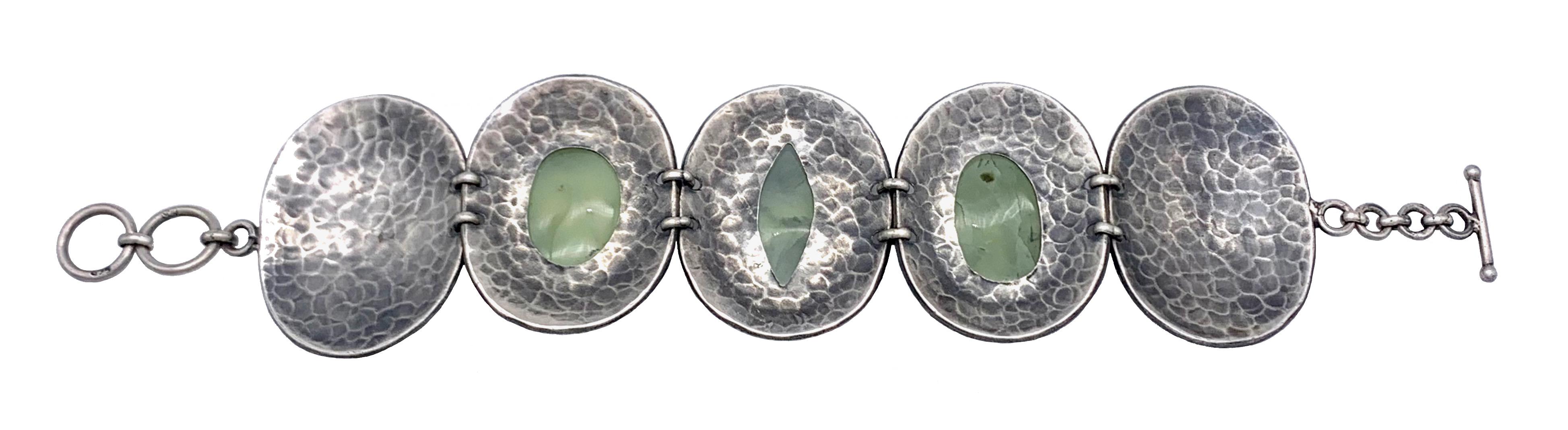 This wonderful chunky silverlink bracelet is set with three unidentified green hardstones. The center stone is the shape of an eye, to either side are two oval stones, all of them in cabochon cut.
The stones are mounted in high, closed settings and