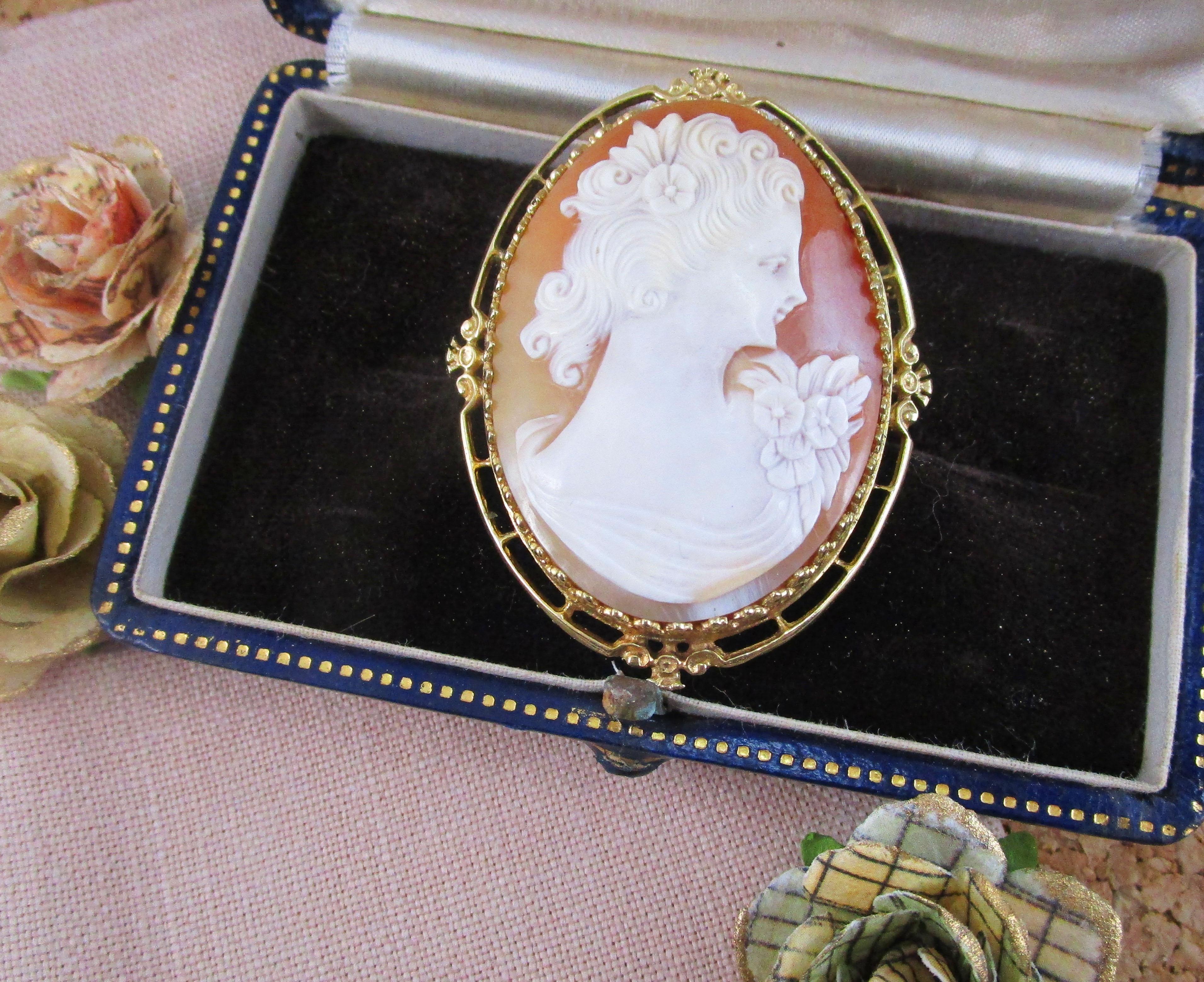 This is a beautiful hand-carved shell cameo from the mid-twentieth century in a delicate 14k yellow gold frame. The cameo itself is delicately carved to depict an elegant woman with fine details in her curling hair and flower adornments meticulously