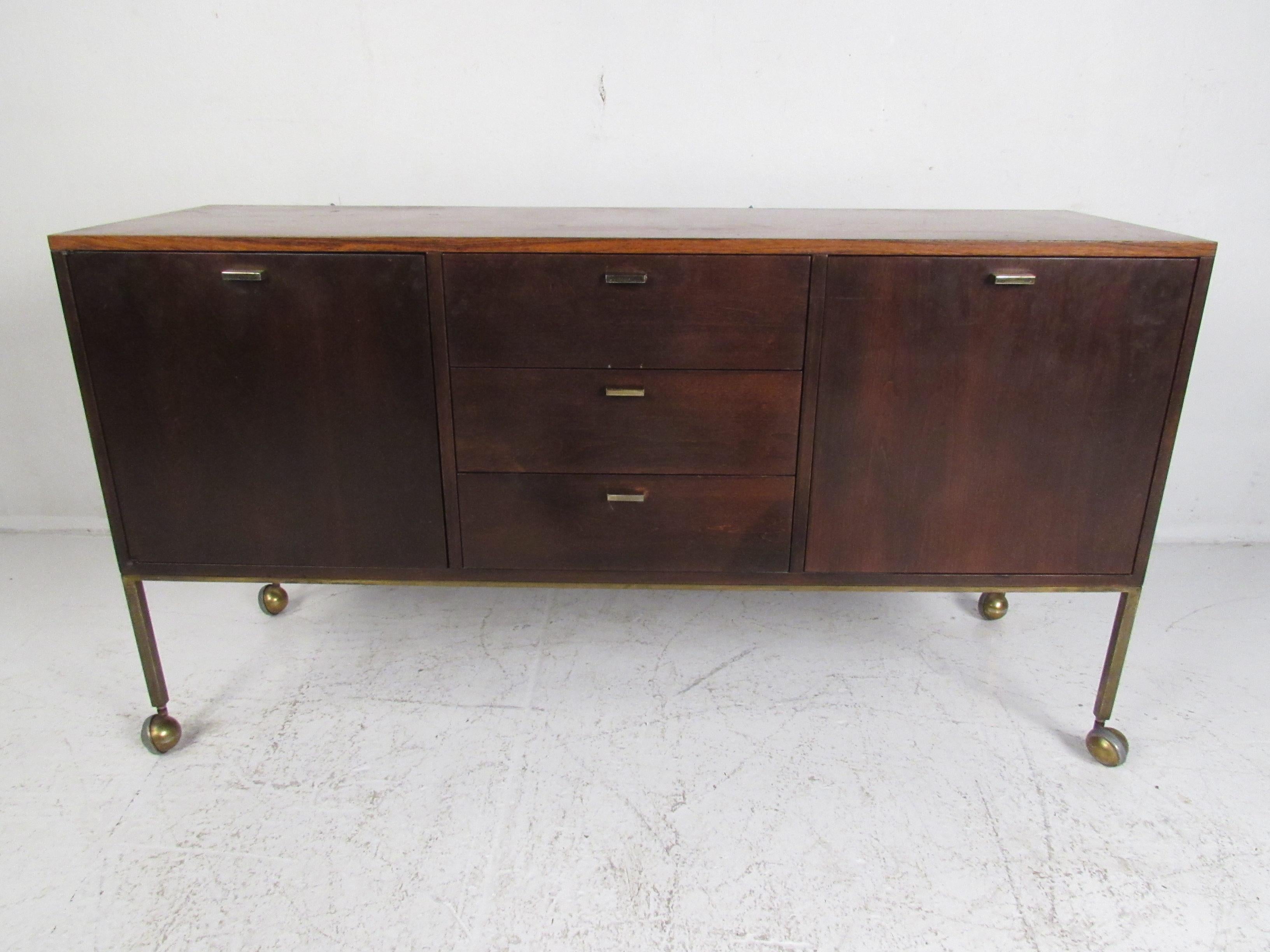 This beautiful vintage modern sideboard boasts a sturdy brass frame with castors ensuring convenient movement. A stunning rosewood wood grain on top adds allure to any setting. This sleek case piece has three hefty center drawers, a large filing