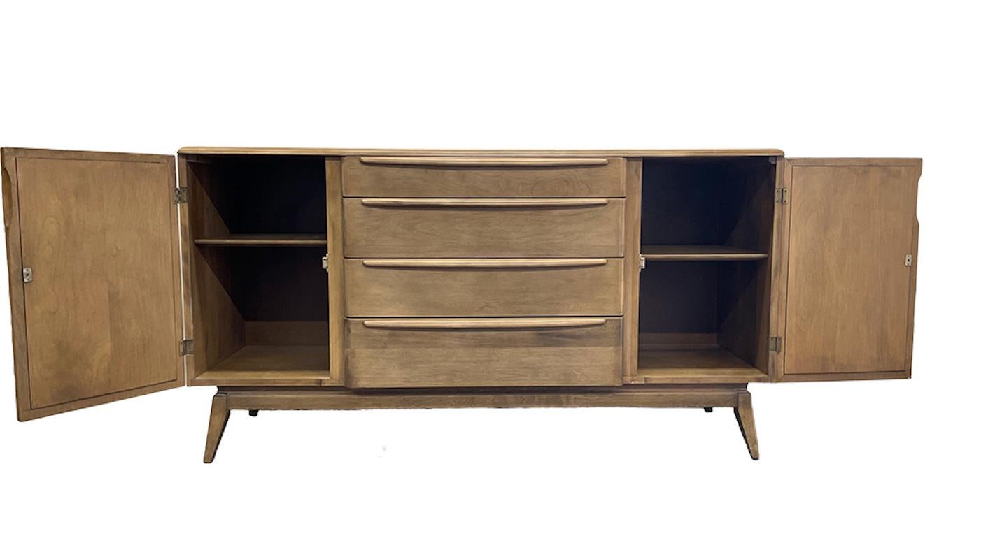 The Heywood-Wakefield Co. buffet cabinet features four drawers including a drawer with removable felt lined silverware organizer that includes the stamp of the maker. the drawers are flanked by two side compartments, each with an adjustable shelf.