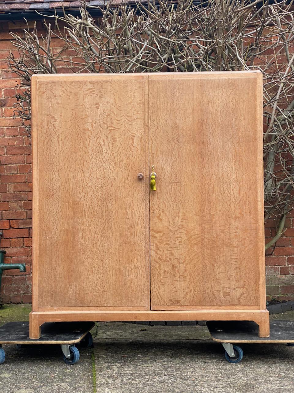 Magnificent early 20th century heal’s attributed limed oak two-door wardrobe compactum, England, circa 1930.

The rectangular top with canted corners over two figured, panelled solid oak doors with carved knob handles opening to reveal brass