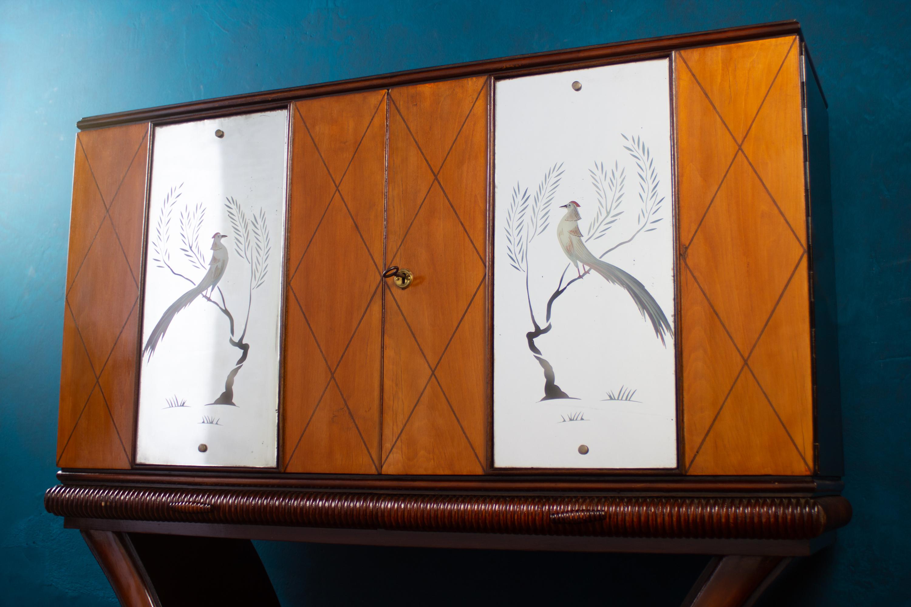 Meuble de bar élégant et inhabituel.
Porte d'entrée décorée de figures d'oiseaux finement gravées. Panneaux de porte à l'intérieur avec miroir en mosaïque et étagères en laiton et en verre. Support avec deux tiroirs coulissants.
Finition bicolore