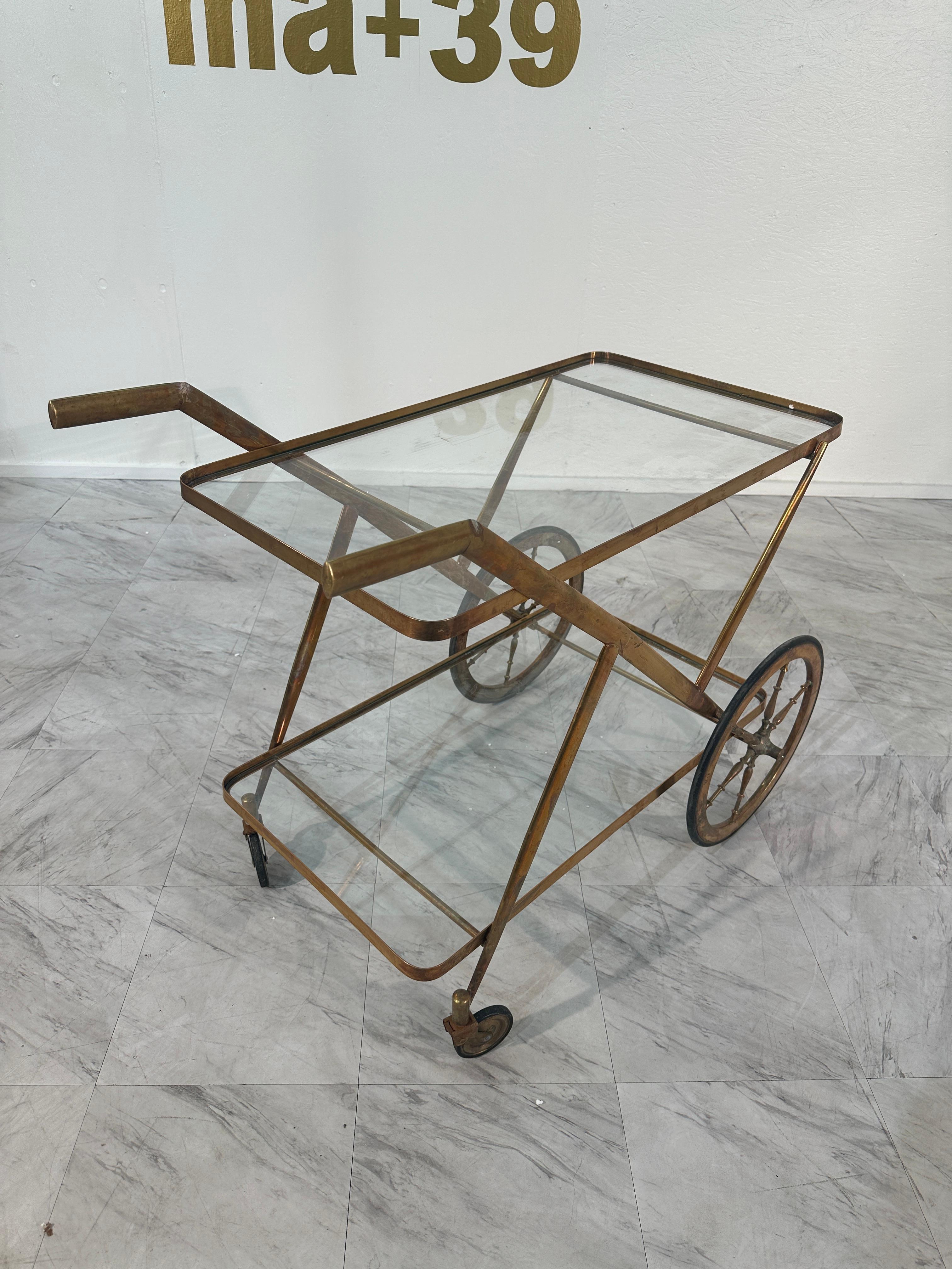 This elegant mid-century bar cart, designed by Cesare Lacca in the 1970s, features a brass frame with sleek black accents and large, stylish wheels. The two-tiered design includes a glass top and bottle holders, making it both a functional and