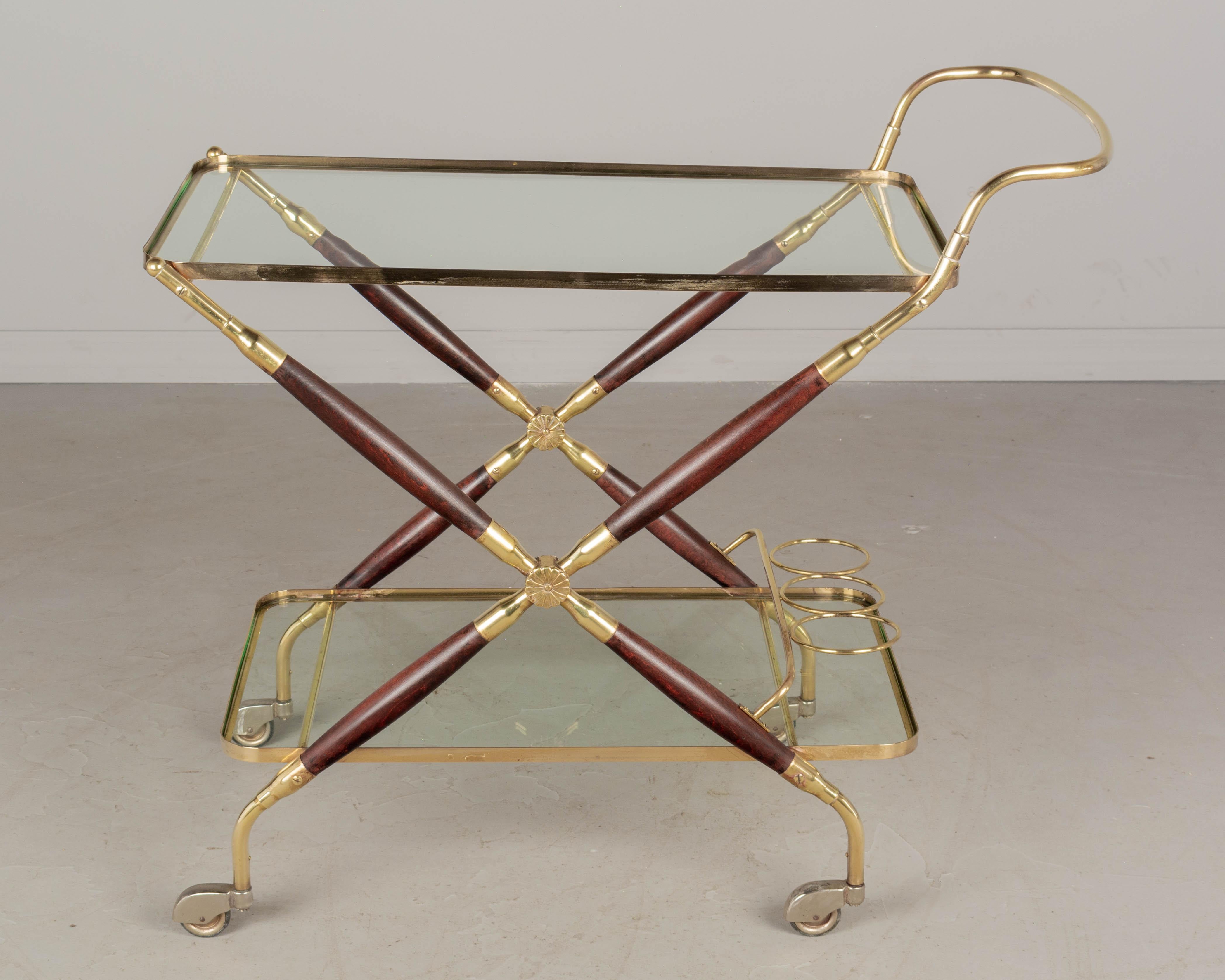 A Mid-Century Modern Italian bar cart by Cesare Lacca. A very nice high quality drinks trolley made of solid brass with turned mahogany stained beechwood x-frame and cast brass rosette details. Glass shelves with a three bottle rack on the bottom.