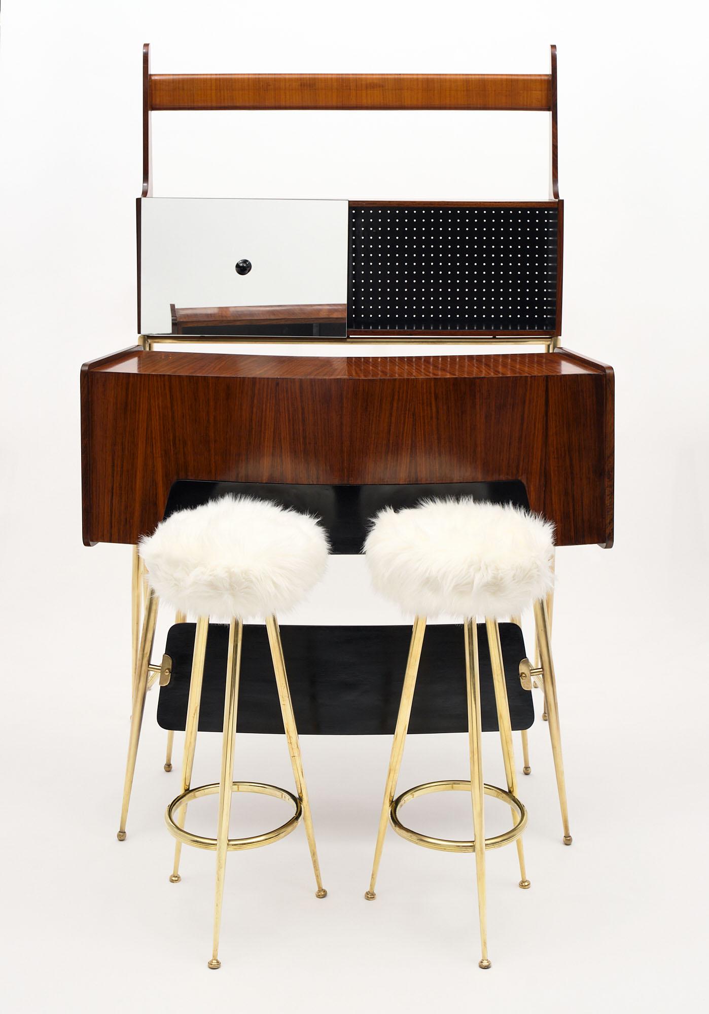 Bar; Italian; made of mahogany and brass. The ensemble features a case on brass legs with a glass shelf and a sliding mirrored door on top; a bar on brass legs of rosewood; and two brass stools upholstered in ivory toned shag fabric. The