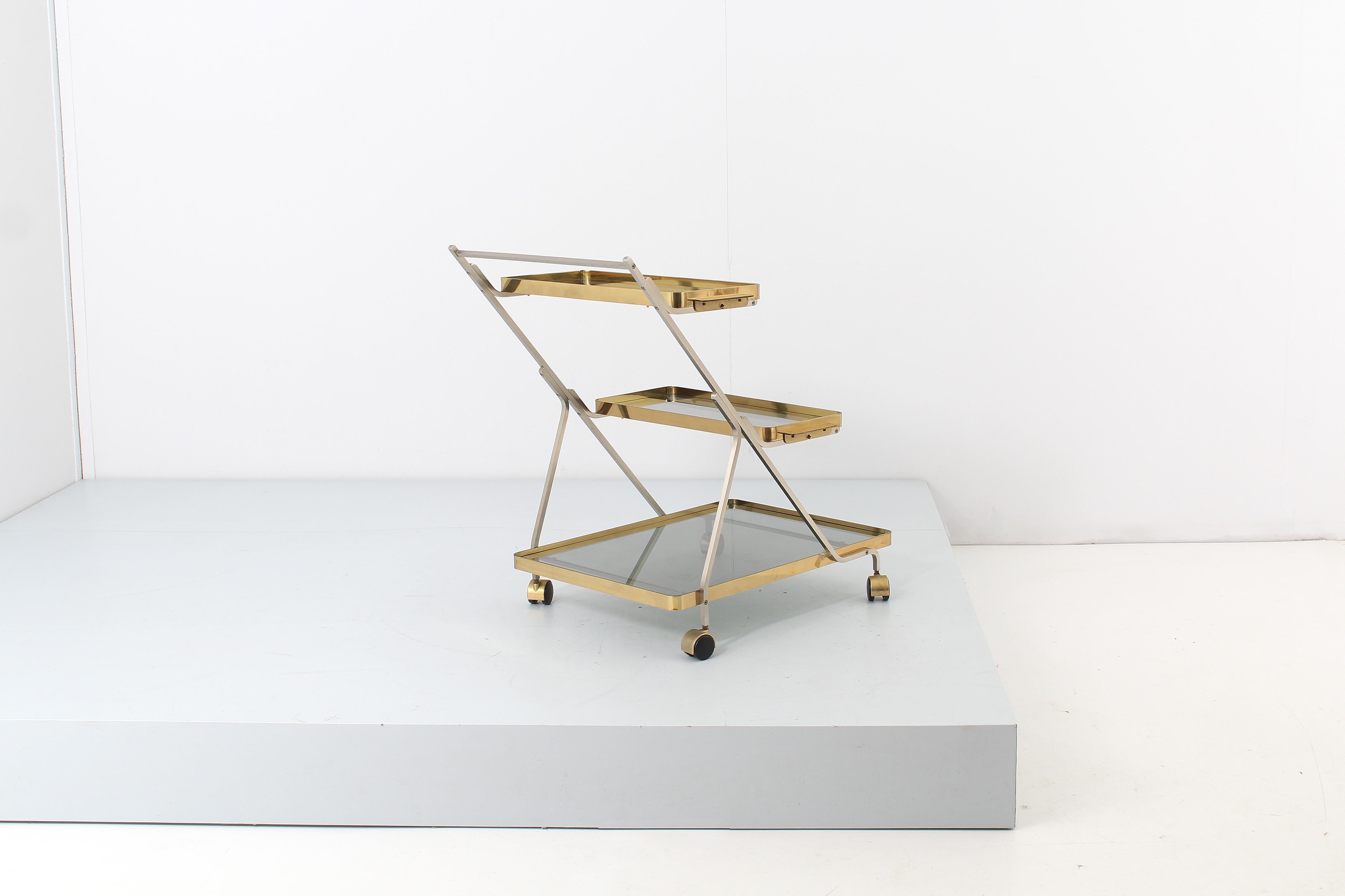 Stylish bar trolley on four wheels, with three shelves with geometric structure in brushed steel and smoked glass shelves with brass support frames. The two narrow trays are removable, with handles obtained on the profiles. Elegant curves of the