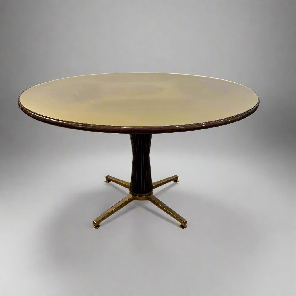 This elegant dining table features an hourglass shaped base of black hardwood with brass inlay. The top is a reverse painted gold tone glass within a beautiful hardwood edging. It rests on four splayed brass feet on casters. 1950's, Italian.