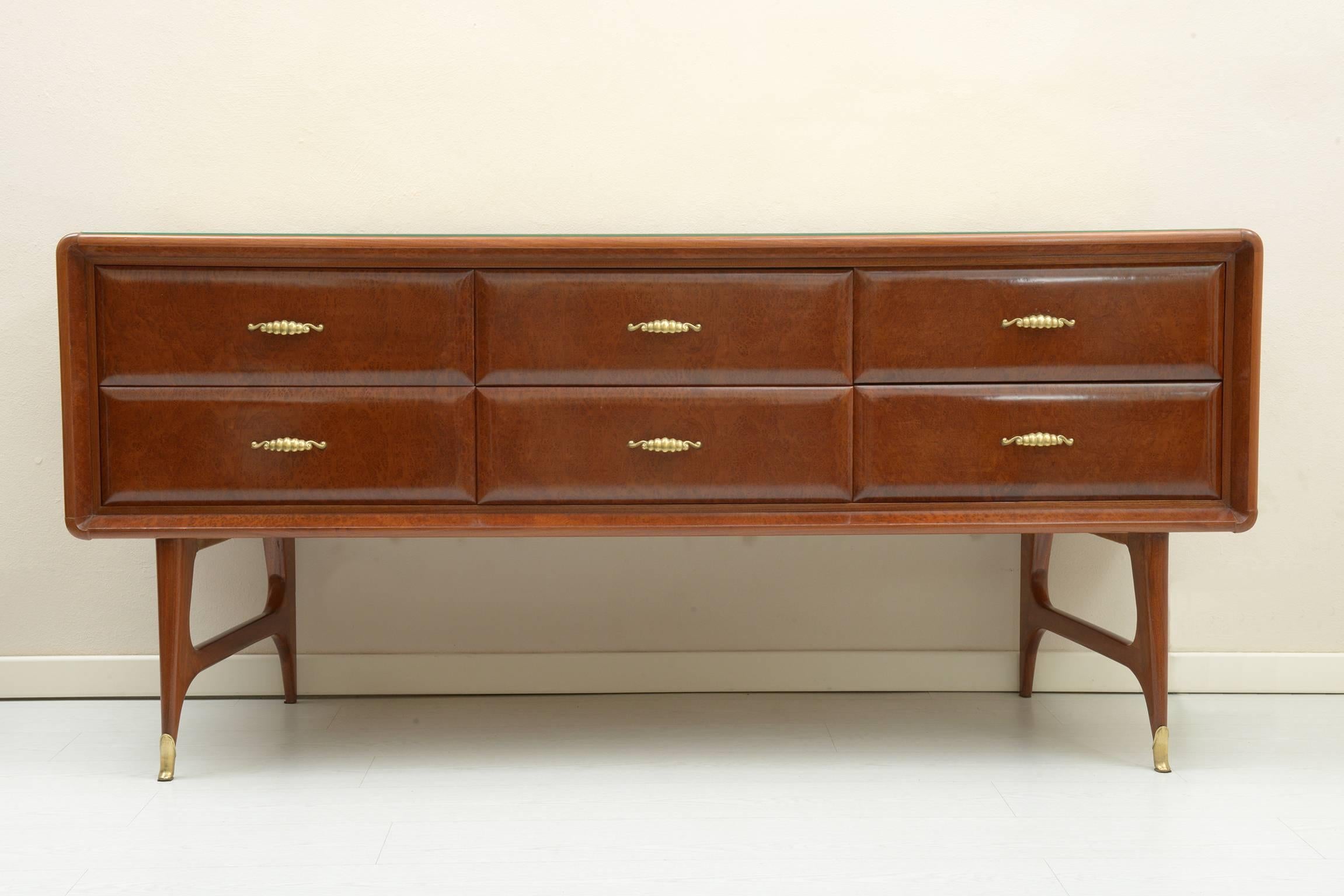 Six front rounded drawers midcentury Italian 1950s chest of drawers on Thuja Burl, cust brass feet and handles.
The lateral support legs are uprights that tilt, squeezing behind towards the central part.
The top glass is gold leaf decorated from