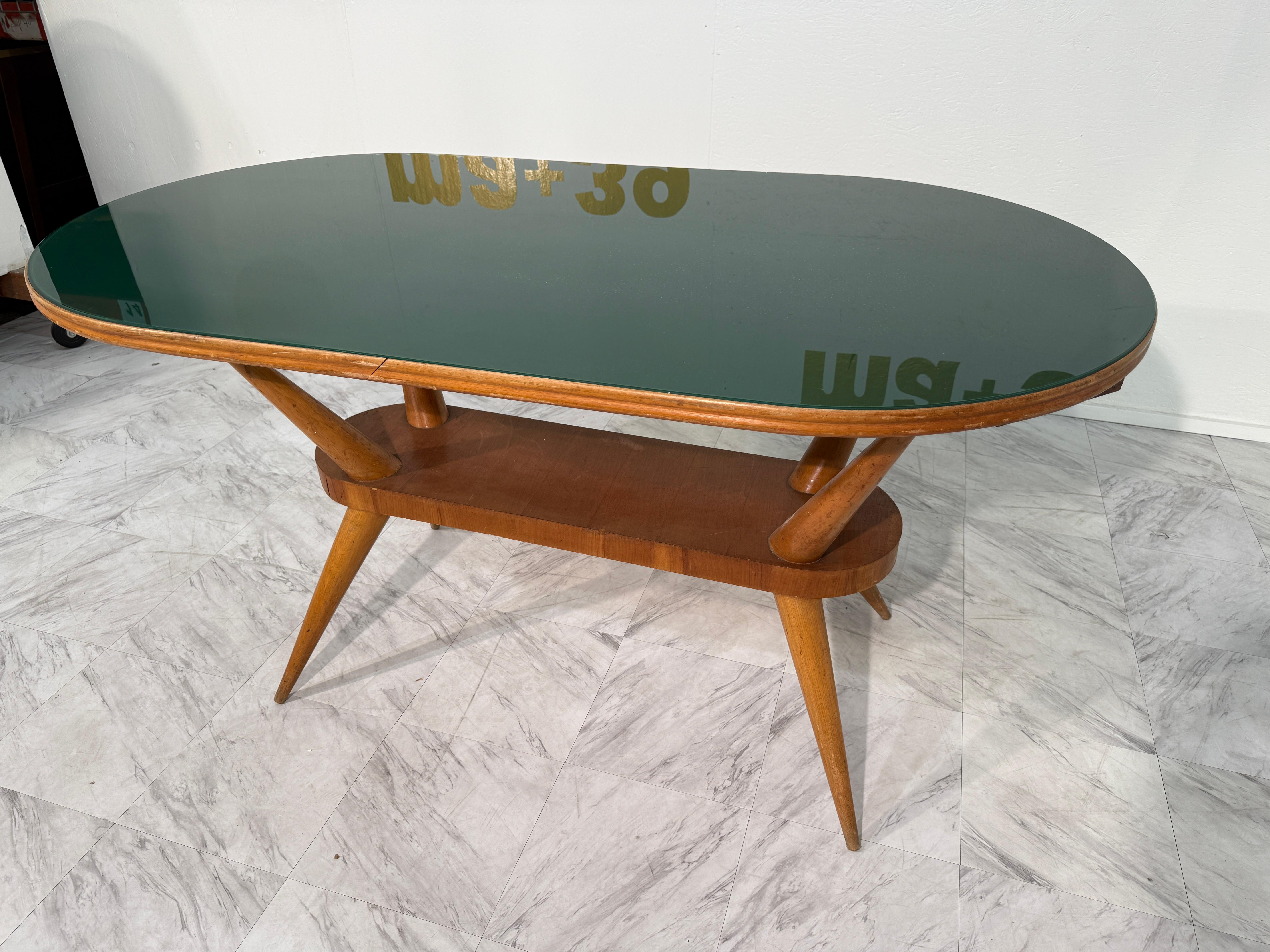 Un tavolino italiano di metà secolo, realizzato negli anni '50, dal design elegante e funzionale. Il tavolo presenta un piano in vetro verde delicatamente ovale incorniciato da un caldo legno color miele e poggia su gambe affusolate e angolate che