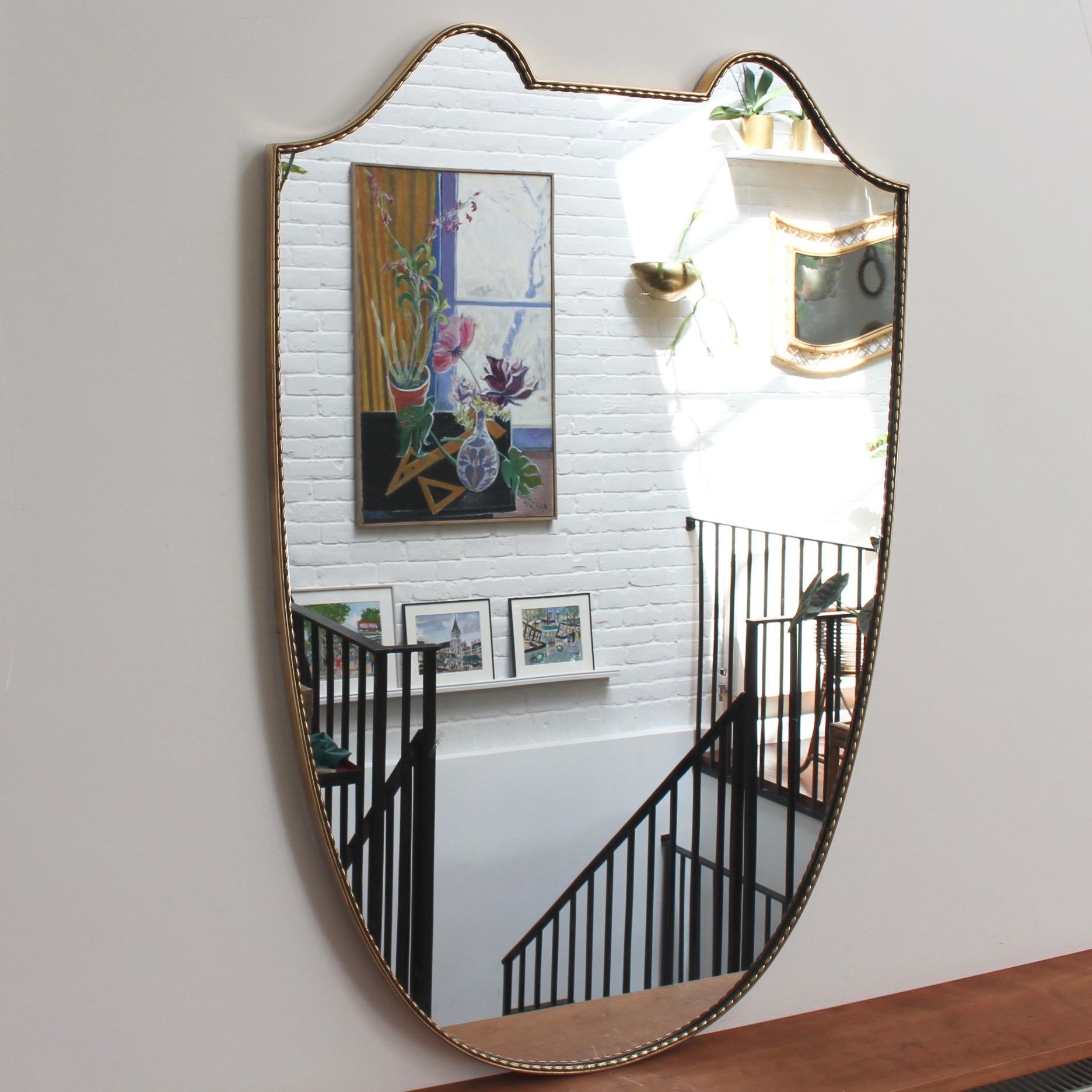 Large Mid-Century Italian wall mirror with brass frame (circa 1950s). This mirror has sensuous curves yet retains its solidity and imposing good looks. The crest-shape is topped by two rounded edges on either side and the frame has a distinctive