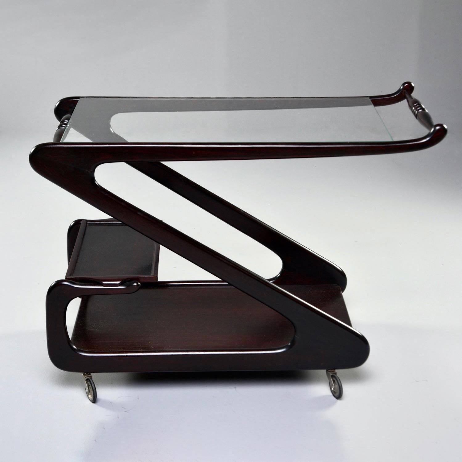 Italian bar cart or drinks trolley has a dark stained wood frame in the shape of a modified Z circa 1960s. Top has glass shelf and handle. Bottom has solid shelf with another smaller shelf that forms a second tier. Unknown maker. Very good vintage