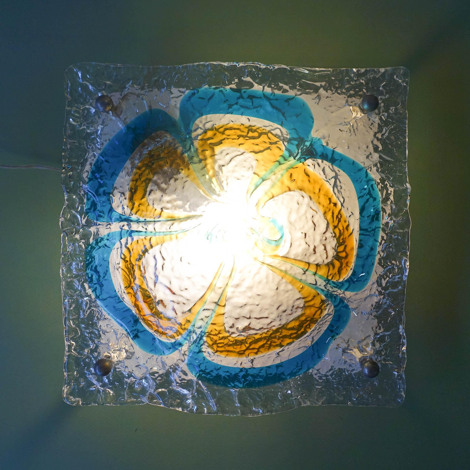 This sconce was designed and produced by Mazzega in Italy, during the 1970's. It features a thick wavy amber, blue and clear Murano glass with a flower-shaped designed, mounted on a white metal wall plate with spherical polished chrome fittings. It