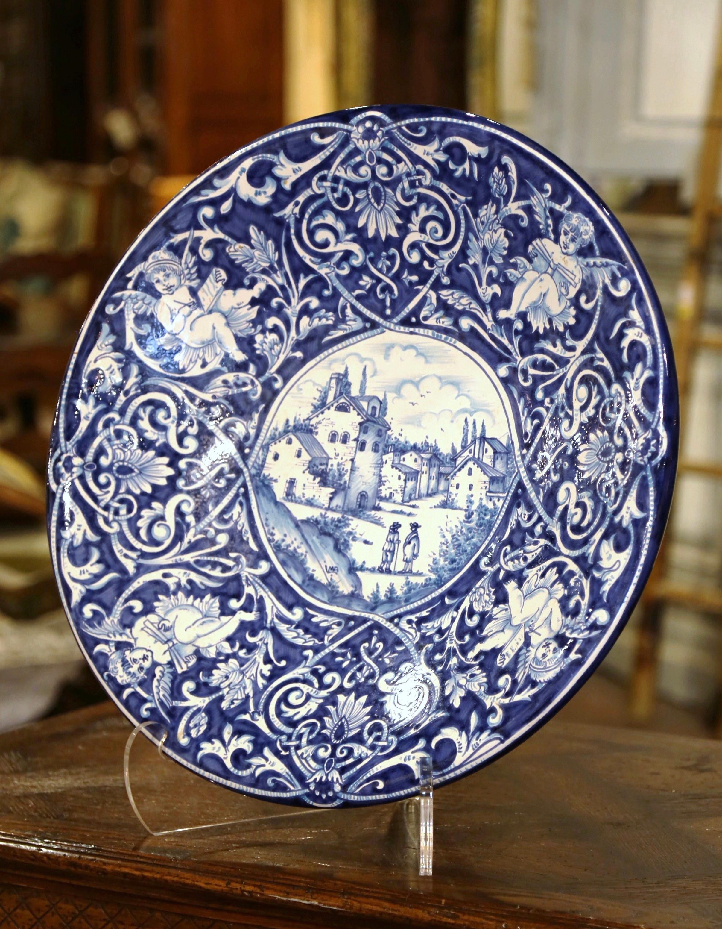 This large antique circular charger was crafted in Italy, circa 1960. The important ceramic wall hanging plate depicts a outdoor pastoral scene with peasants talking in a village with farmhouses around. The border is further embellished with cherub
