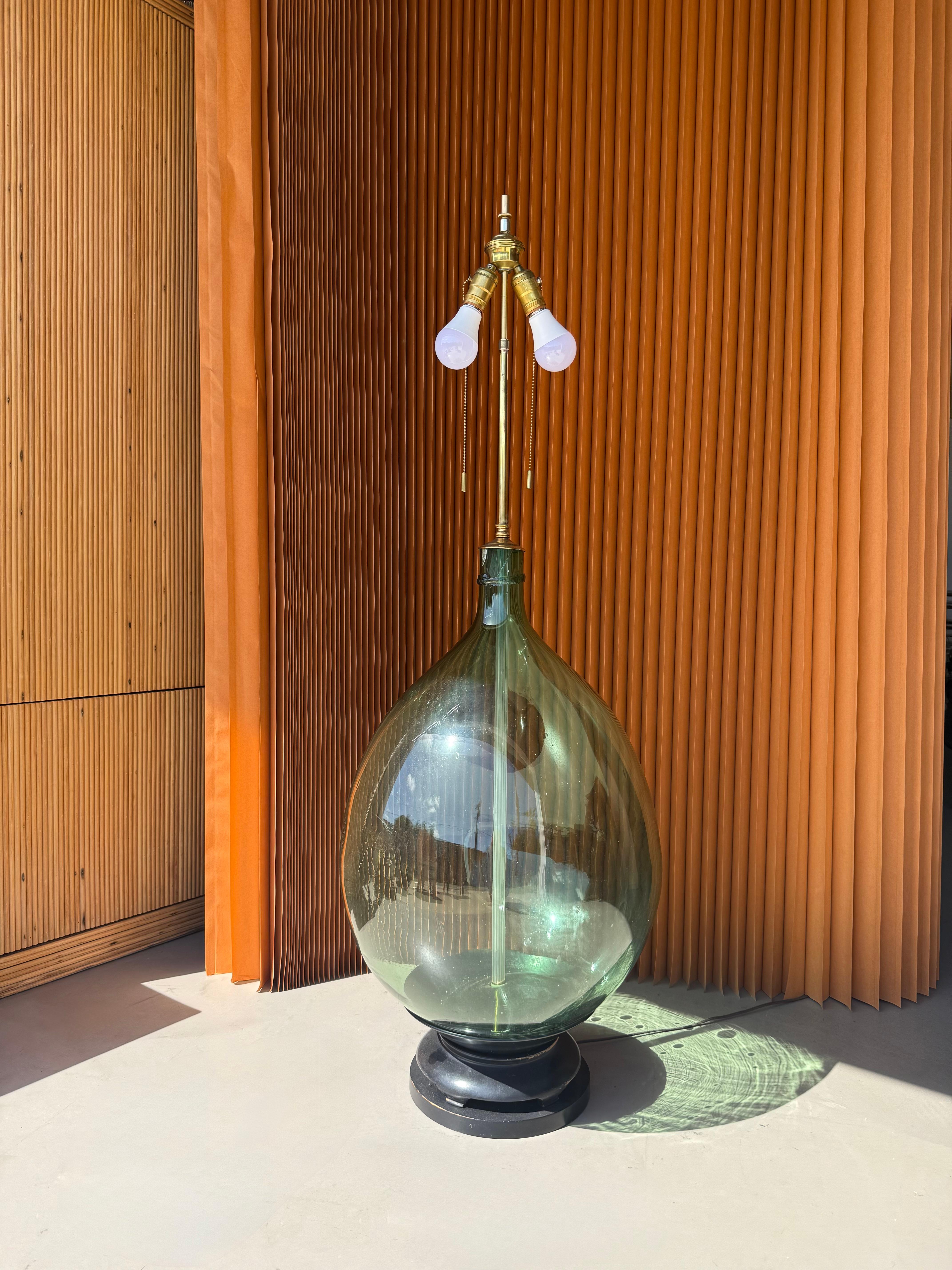A stunning mid-century Italian table lamp repurposed from a hand-blown fiasco demijohn bottle, featuring a classic bulbous silhouette in subtly tinted green glass. The original glass vessel, traditionally used for wine or oil, is mounted on a round