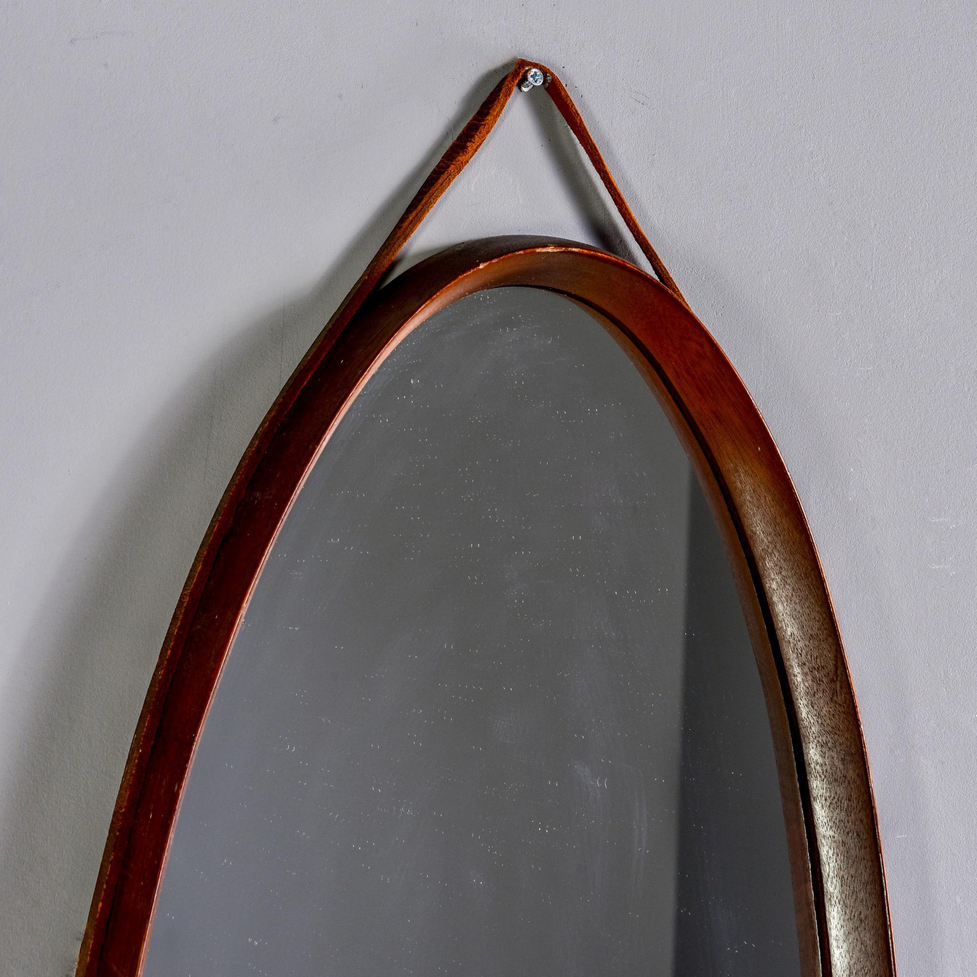 Circa 1970s Italian mirror is an elongated oval with deep set mahogany frame and leather hanging strap. Unknown maker.