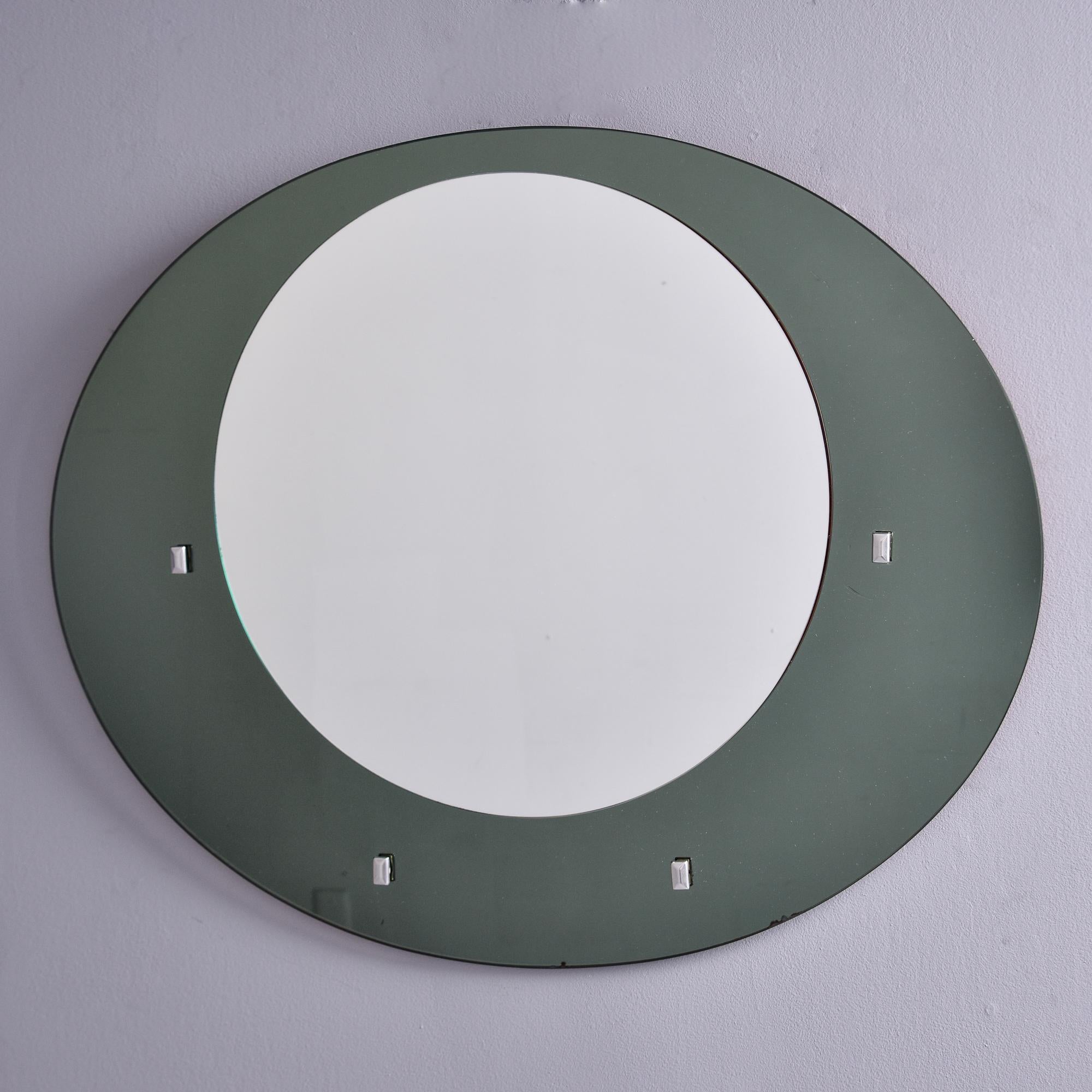 Found in Italy, this round mirror with gray glass frame dates from the late 1960s. Round mirror is set off-center on top of a gray glass oval frame. Rectangular silver tone metal accents. Metal hanger on back. This mirror is in the style of Fontana