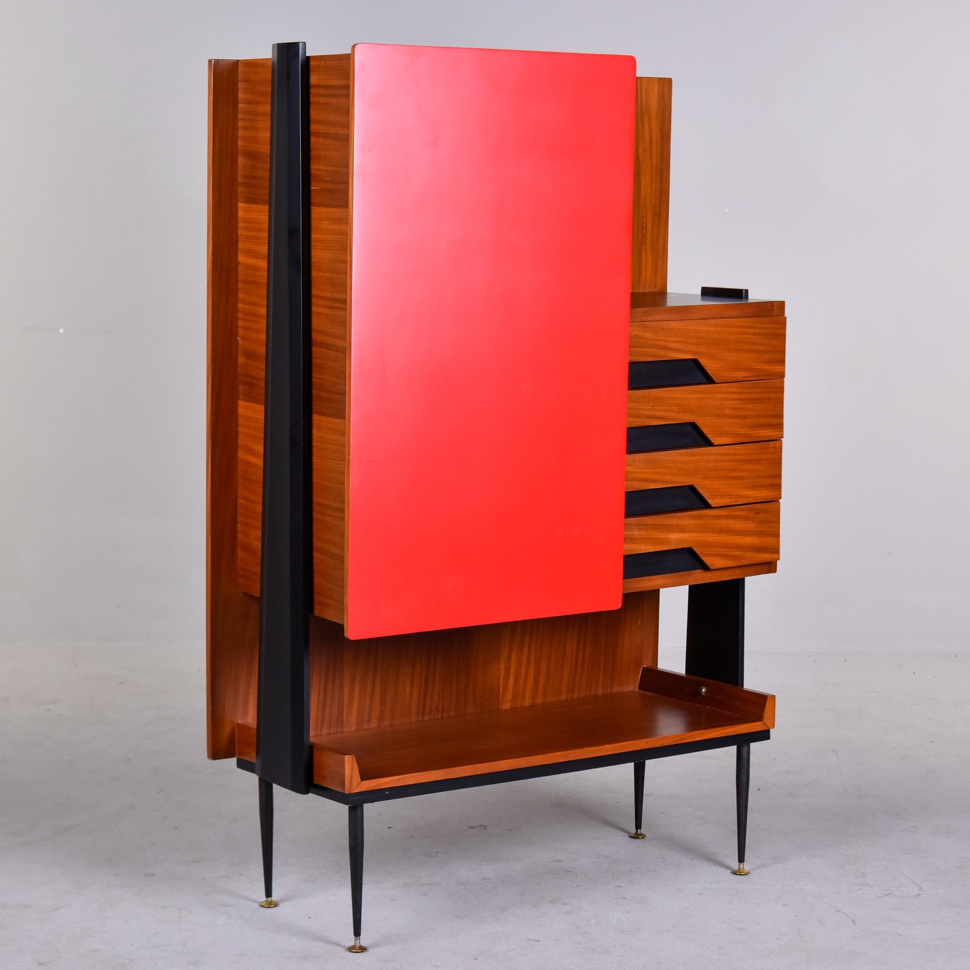 Found in Italy, this tall cabinet dates from the 1960s. Teak base with contrasting black side supports and a bold, red cabinet door. Narrow black metal legs with brass feet with an open lower shelf, four functional drawers with dovetail
