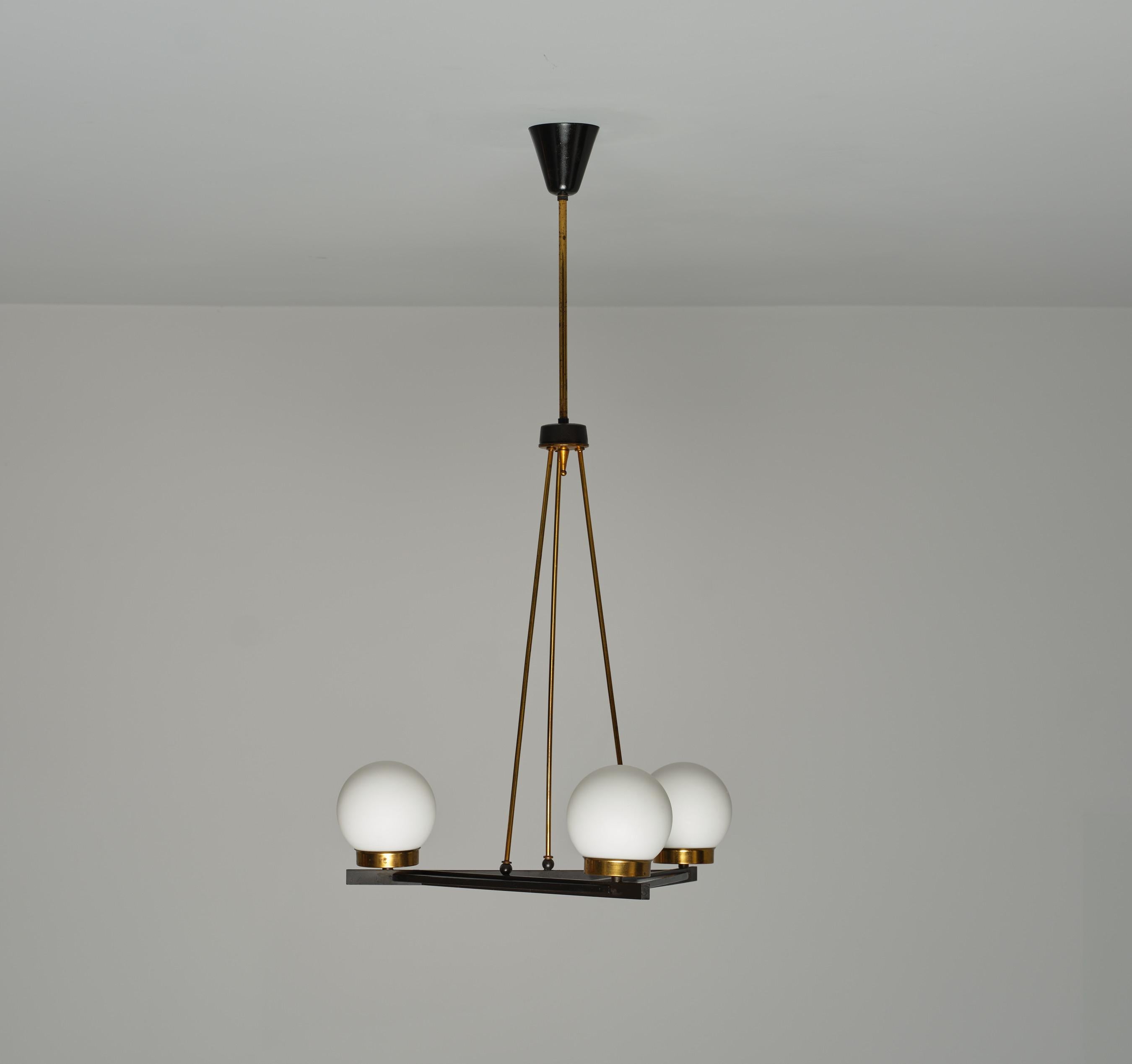 This Italian mid-century pendant light features three opaline glass globes as shades, each mounted on a triangular brass frame with its original patina, combined with black lacquered steel and lacquered wood accents. The fixture is equipped with