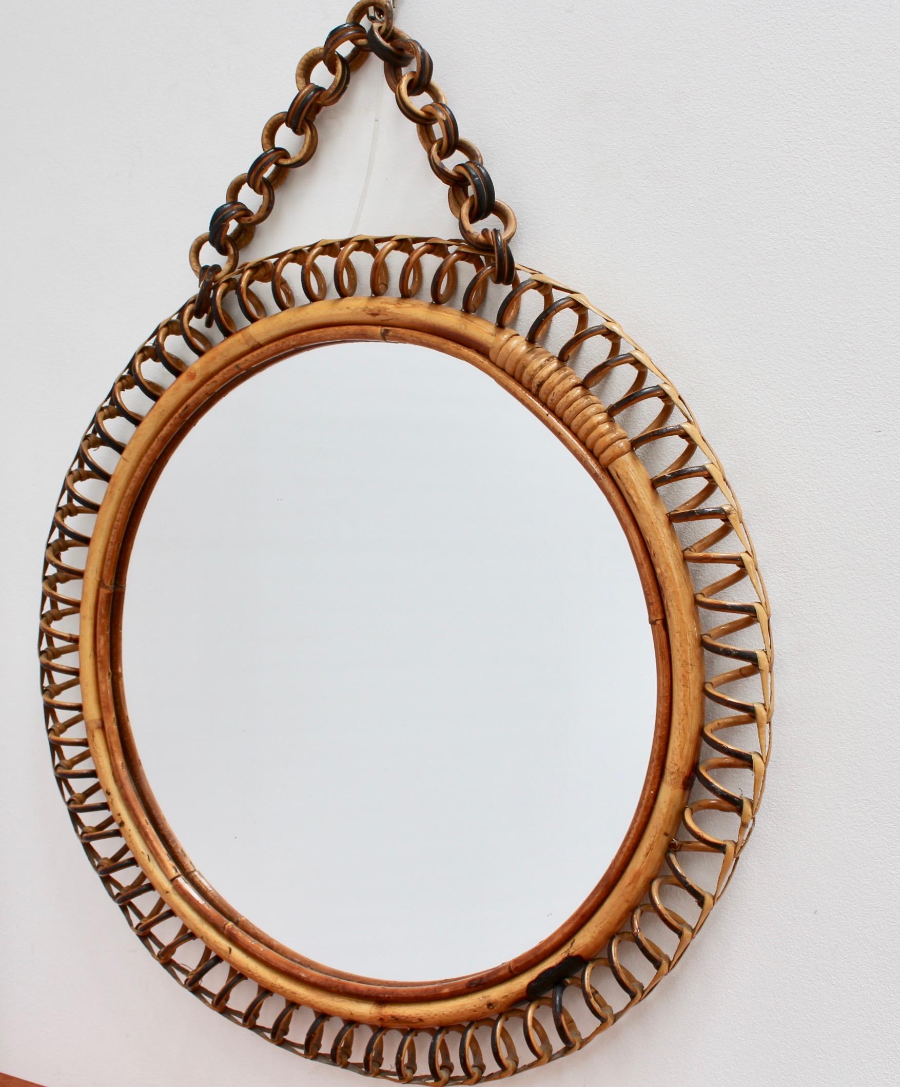 Italian circular-shaped rattan wall mirror (circa 1960s). This mirror has a very charming round shape with rattan spirals framing the mirror. The rattan hanging chain adds further allure. There is a characterful, aged patina on the mirror frame. In