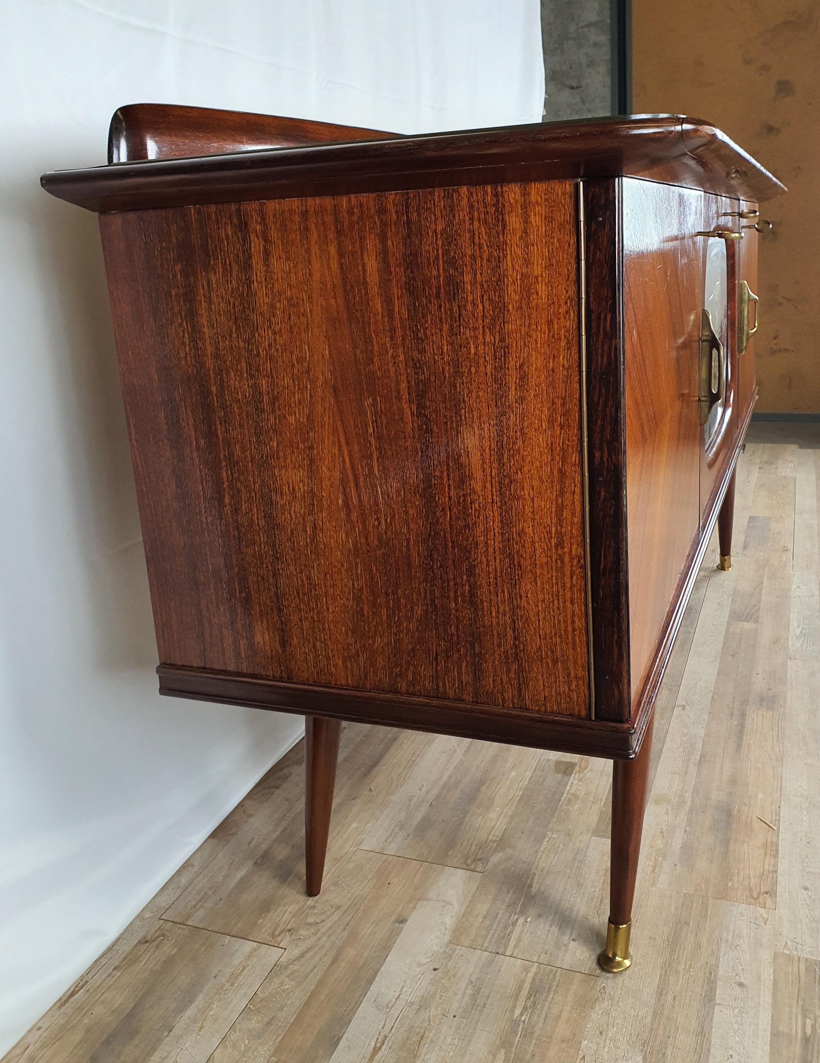 Elegant early 1950s hall or living room sideboard in walnut with brass details and a flap with bar area in the centre.

It is a piece of furniture with a Vintage Design but at the same time with a touch of modernity given by the sinuous and soft