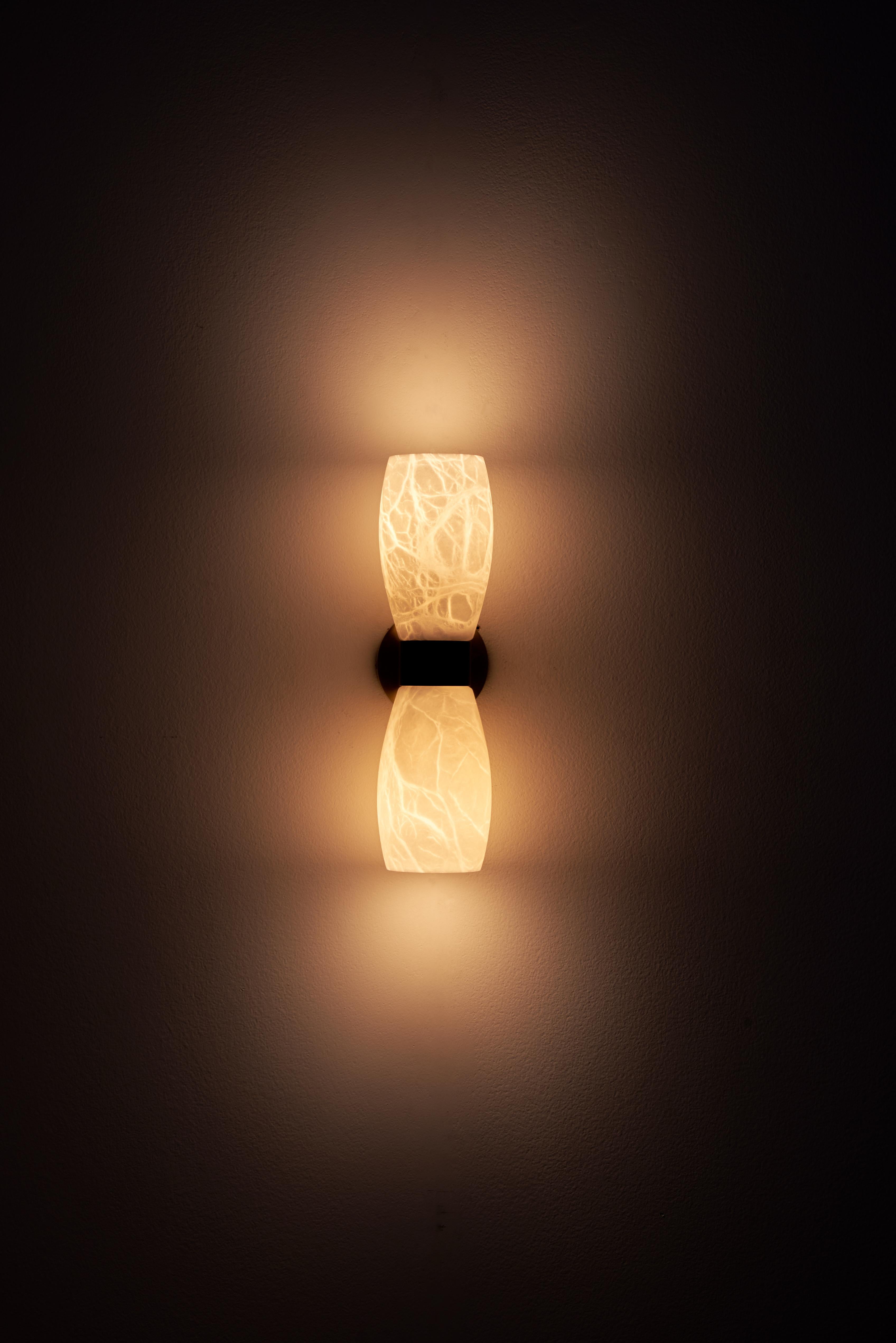 The Demetra wall sconce, inspired by the mid-century style, combines French gold and matte black finishes with alabaster to create a mesmerizing display.
The ethereal glow emitted by the illuminated cups seems to hover in the air, casting a gentle,