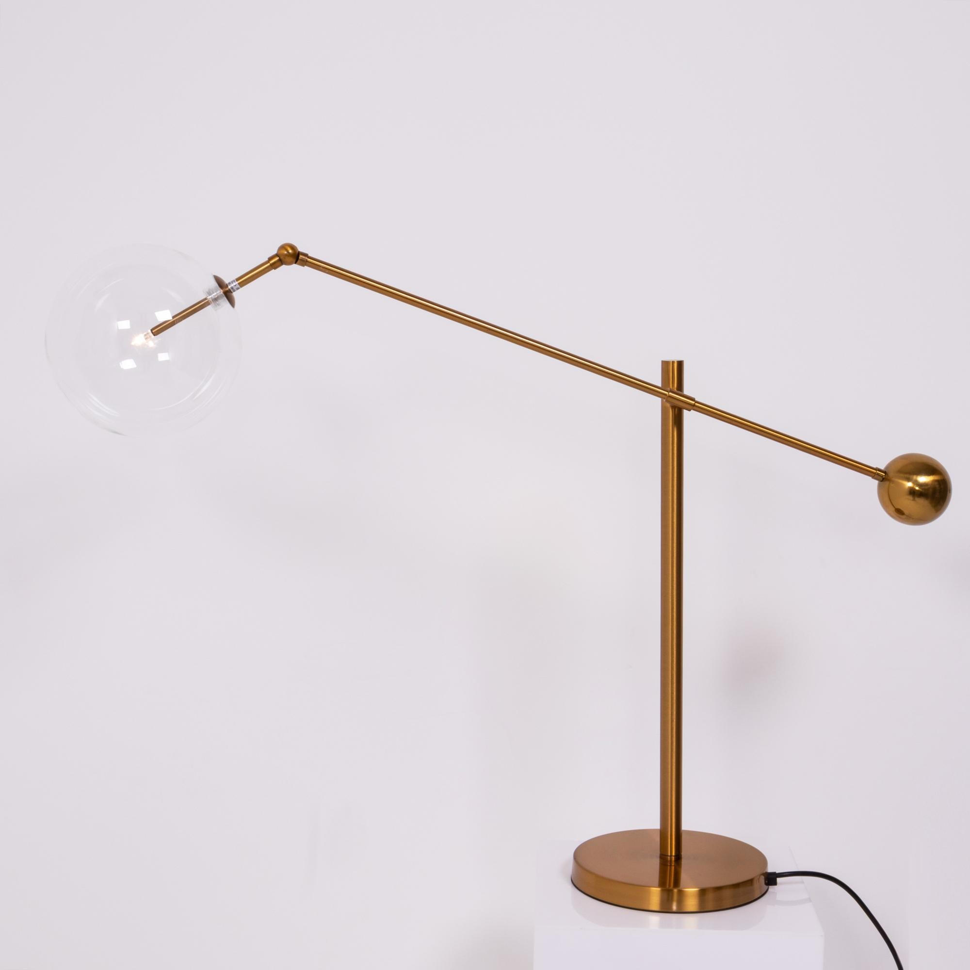 Inspired by mid century Italian design, this table lamp features a striking globe shade in clear glass.

Sitting on a round brass base, the arm cantilevers off of the stand and is weighted at the end with a brass sphere.

The lamp has a UK plug