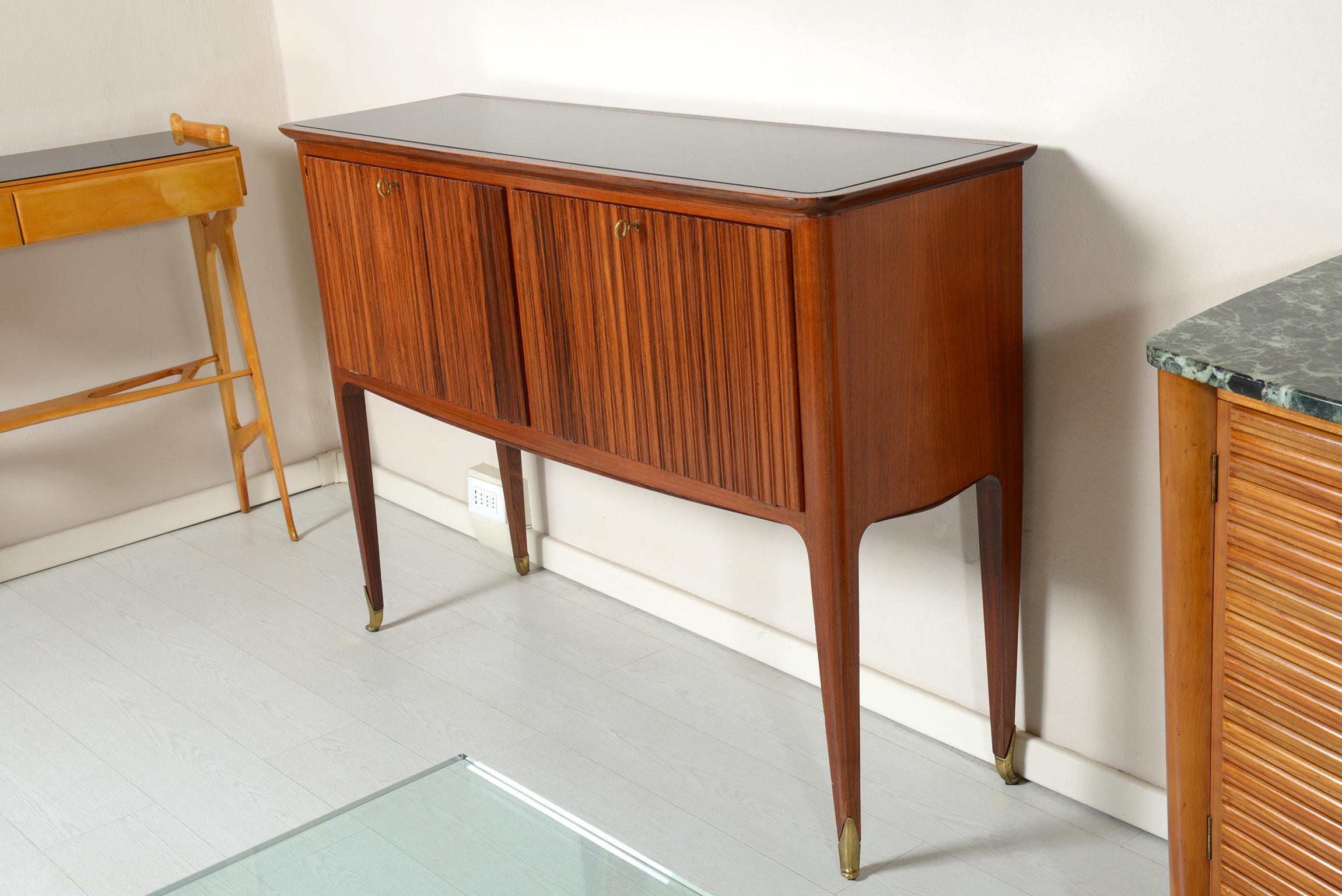 Italian sideboard in walnut grooved body, four slender legs ending with cast brass feet.
Two-flap doors with shelves inside.
Recessed black glass top.
Mid-Century Modern.
           