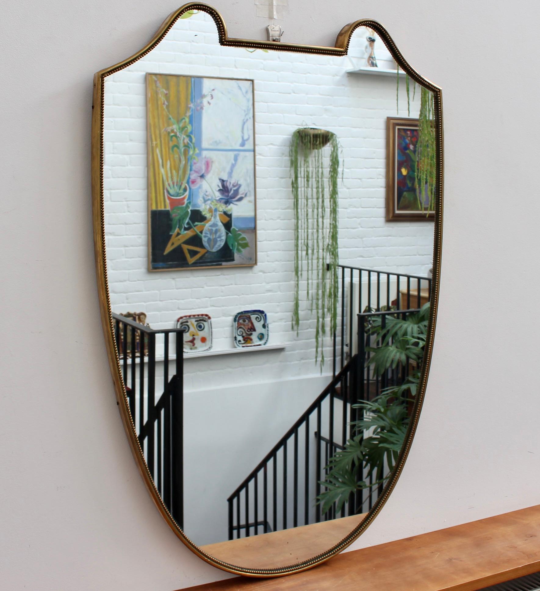 Vintage Italian wall mirror with brass frame (circa 1950s). The crest-shaped mirror is substantial with beading and sensuous curves, like ears, crowning the piece. It exudes character, solidity, weightiness and has eye-catching good looks. The two