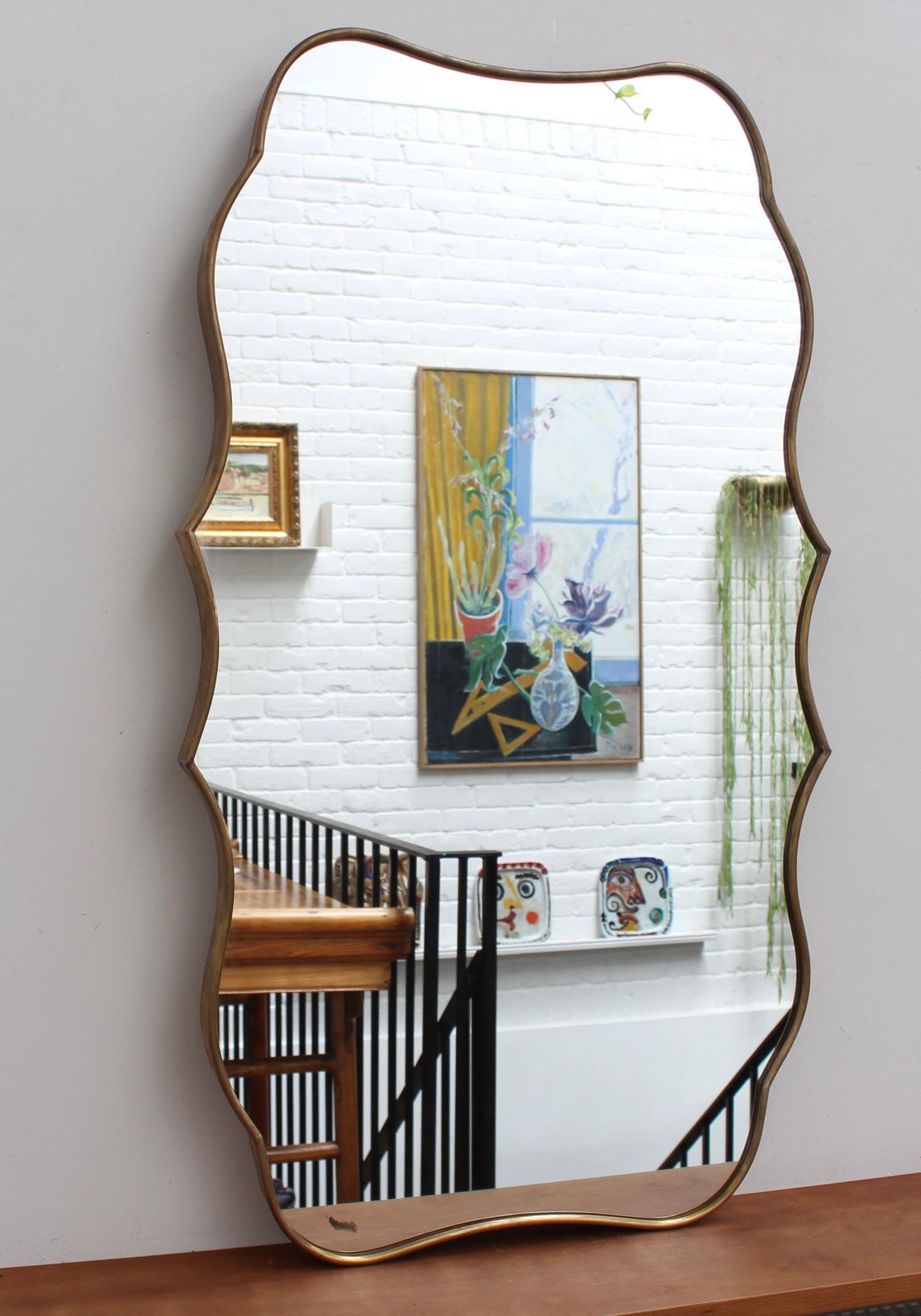 Italienischer Wandspiegel aus der Mitte des Jahrhunderts mit Messingrahmen (ca. 1950). Dieser Spiegel hat eine etwas ungewöhnliche, aber einladende Form - durchgängig elegante Kurven und deutliche Punkte sowie eine massive Holzrückwand. Sie hat
