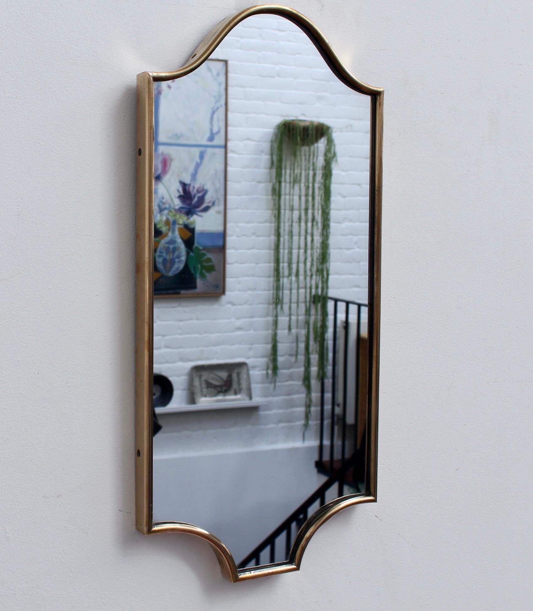 Small-scale midcentury Italian wall mirror with brass frame (circa 1950s). The mirror is rectangular-shaped with a sensuous curve on top and on the bottom, two curves connecting to a short horizontal. It is classically elegant and distinctive in a