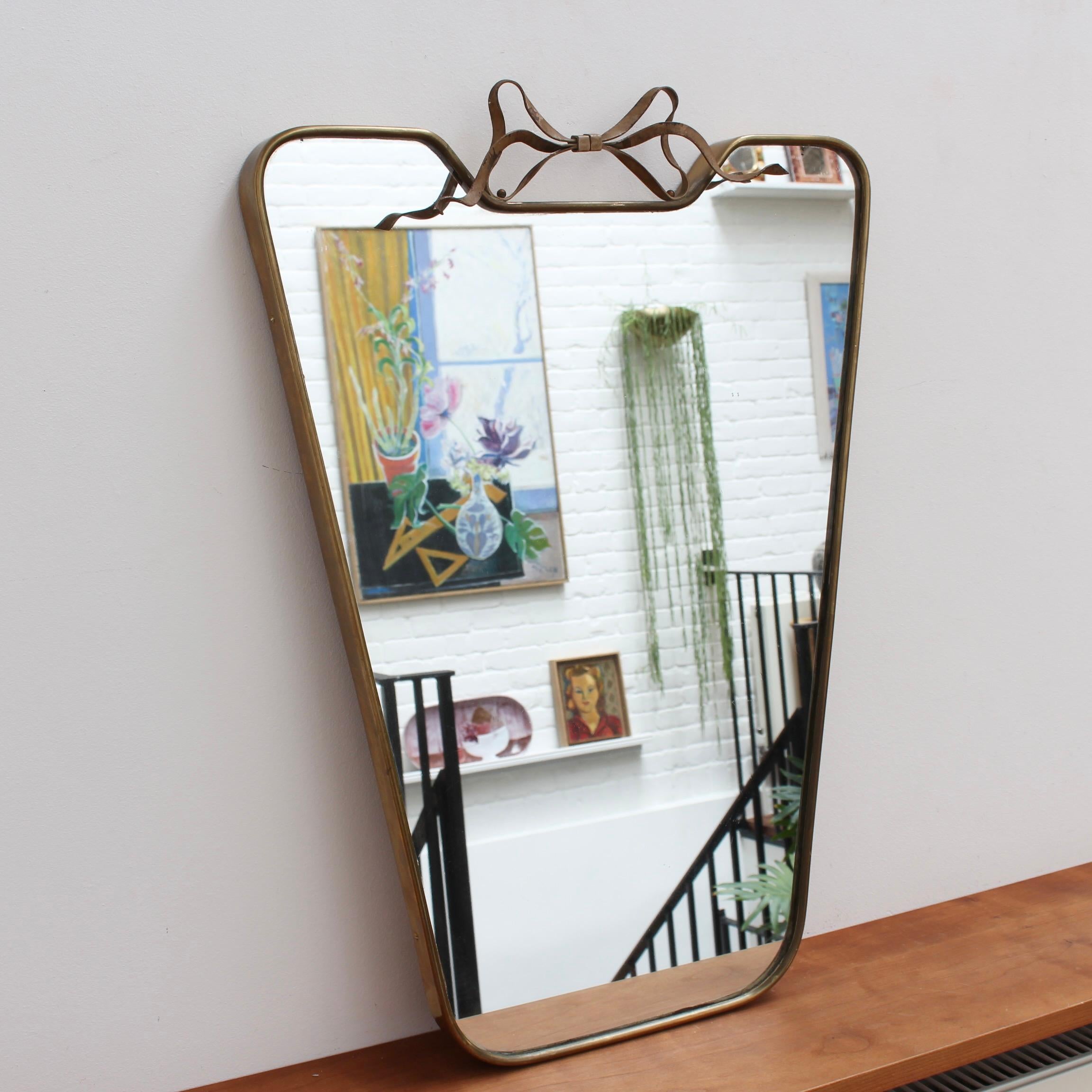 Mid-century Italian wall mirror with brass frame and top flourish (circa 1950s). This mirror is simply elegant and characterful in a modern, Gio Ponti style. It is classically-shaped presenting a charming top flourish in the form of a ribbon. The