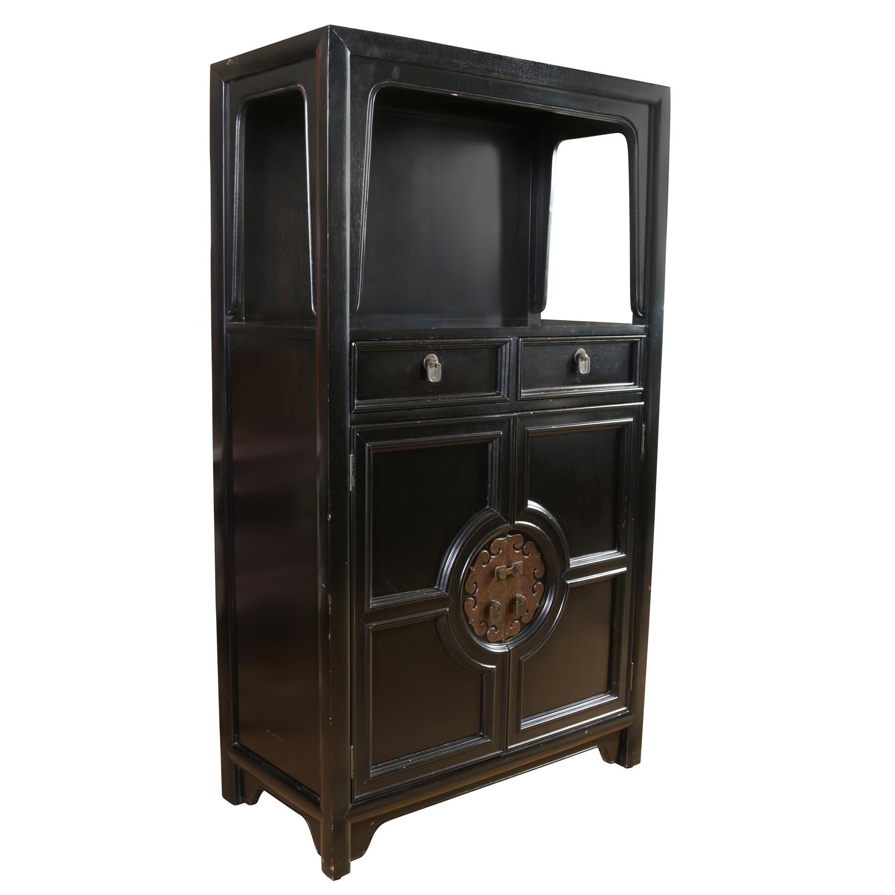 A James Mont style midcentury black wood cabinet with vintage Asian Style hardware. The cabinet has an open shelf at the top bordered by a shaped apron, two small drawers with brass hardware and then two doors at bottom that open to reveal three