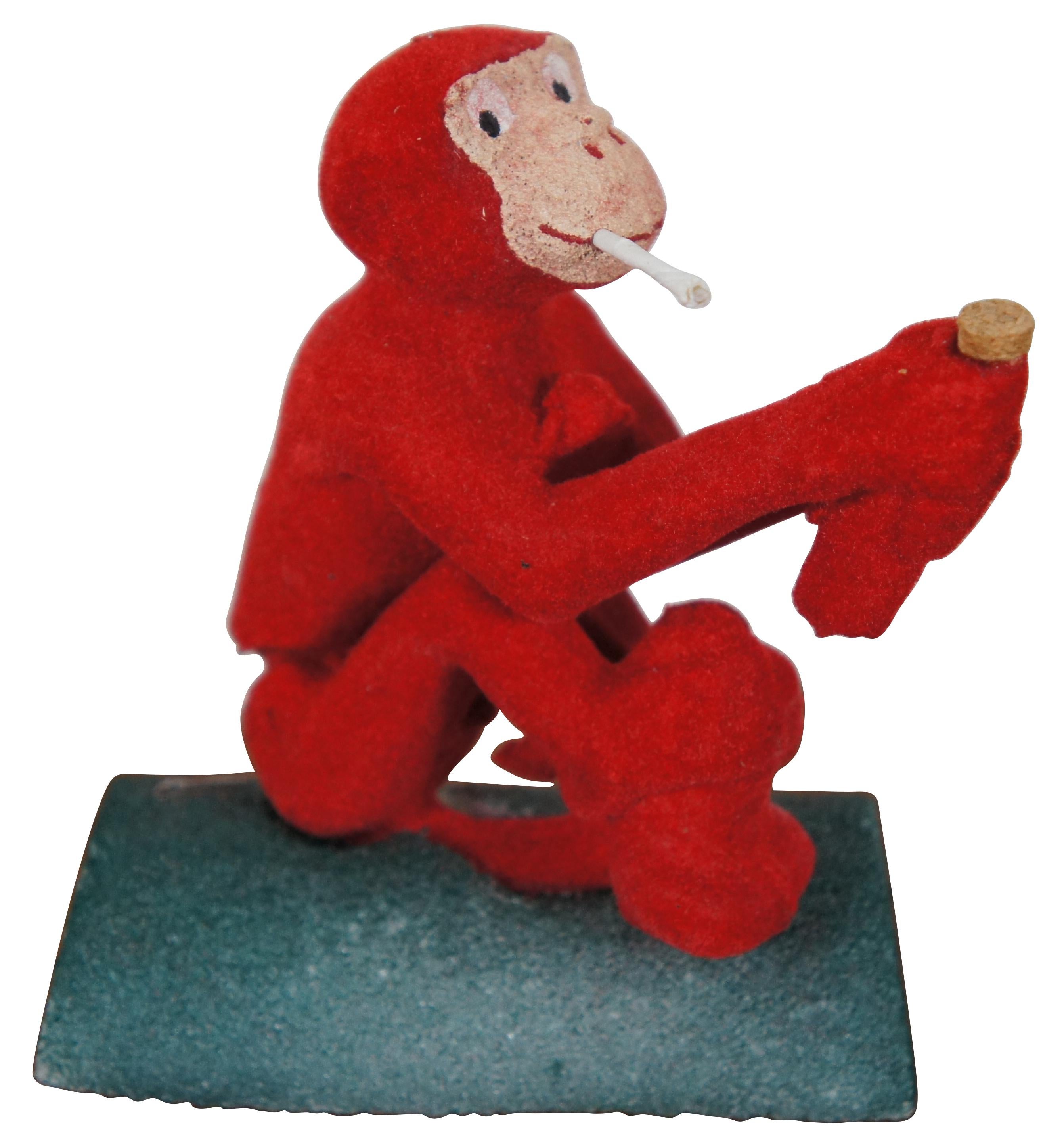 Rare 1950s novelty smoking monkey toy with velvety red fur and painted face, set firmly in green painted styrofoam. Monkey hinges back at the waist and holds a bottle in his hands. Hole in smile accommodates the self-smoking Smokie “cigarettes” (one