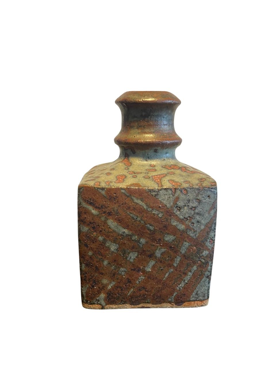 Un étonnant vase bouteille en poterie d'atelier japonaise, datant probablement du milieu du 20e siècle, qui témoigne d'un savoir-faire magistral et d'un jeu unique de textures et de glaçures. La pièce présente une base carrée distinctive qui se