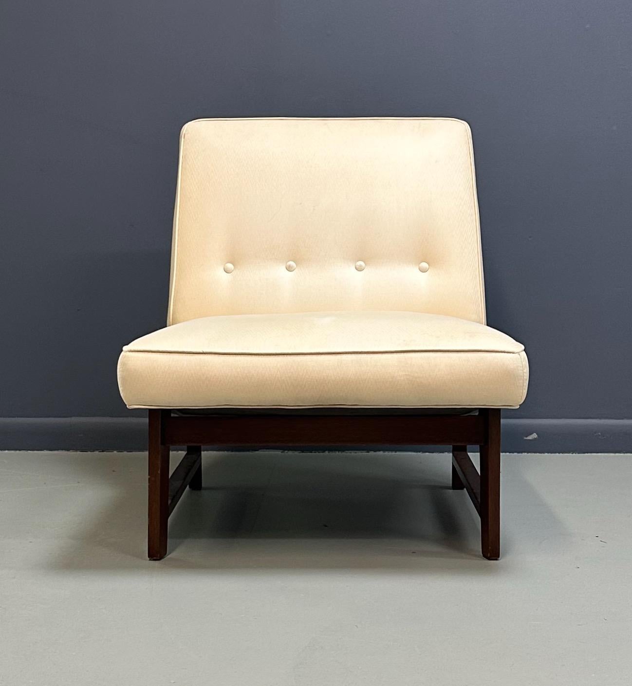 Una refinada silla tipo zapatilla moderna de mediados de siglo al estilo de Jens Risom, con un perfil bajo y una elegancia discreta. La silla está tapizada en un suave tejido crema con un sutil mechón de botones en el respaldo suavemente inclinado,