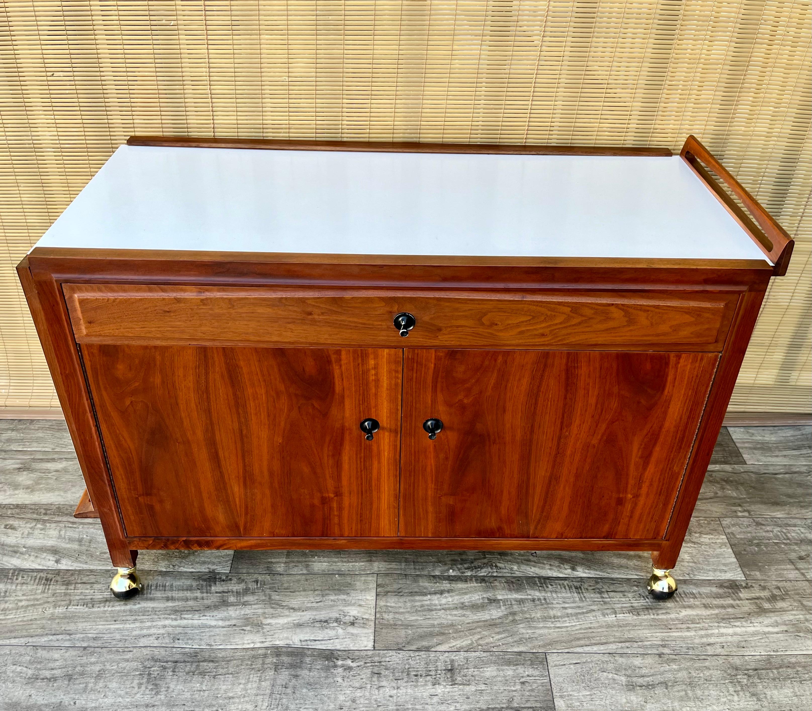 Vintage Mid Century John Stuart Buffet Server / Bar Cart. Circa 1960s. 
Features a quintessential sleek Mid Century Modern Design, two side cabinets, a top drawer, a expandable white laminated top, finished back, and casters for easy mobility. 
In
