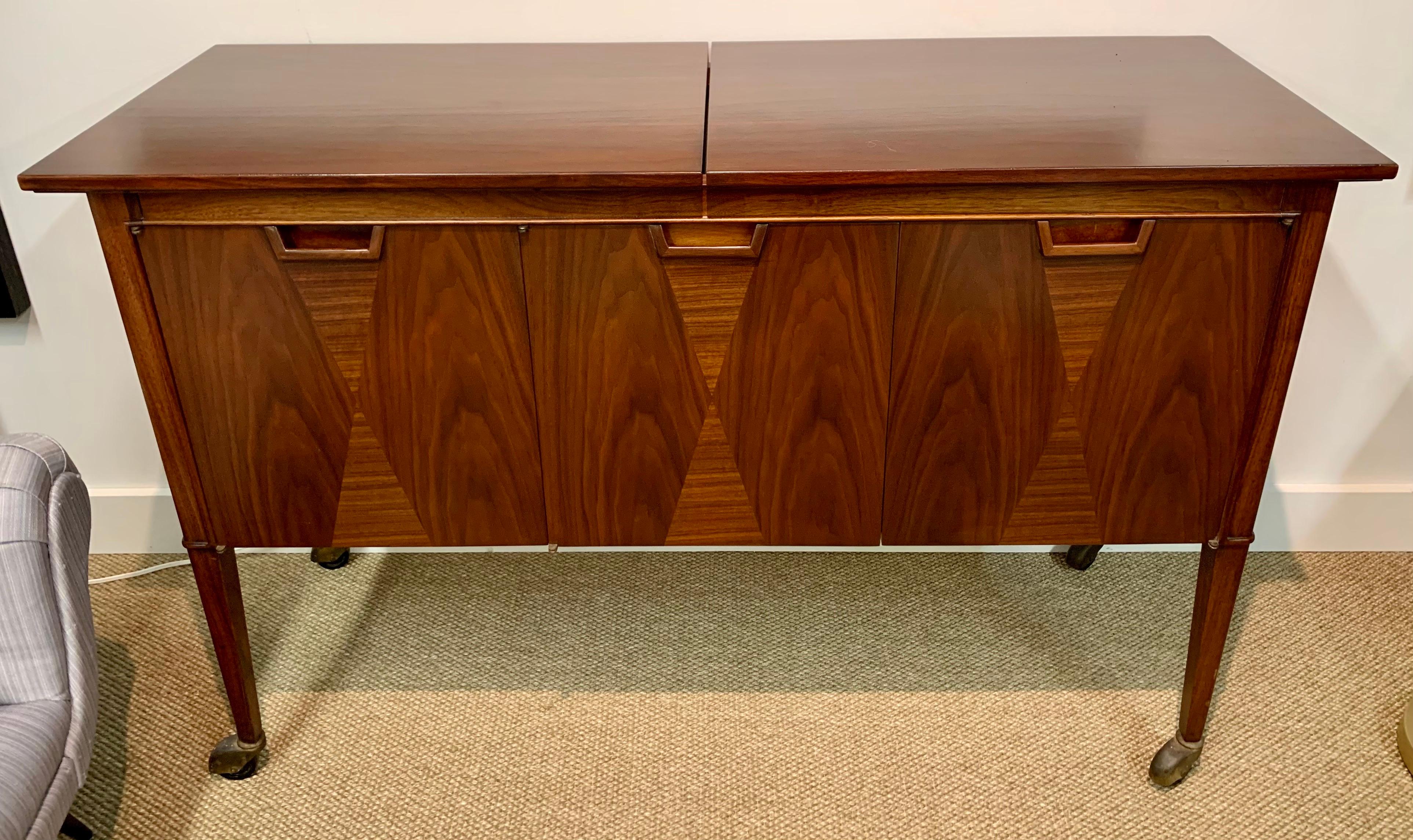 Beautifully restored John Stuart for Janus Collection midcentury rolling dry bar or buffet features beautiful walnut graining and accented by hourglass shaped alternating veneers. The top expands to reveal a black formica top with raised edges. The