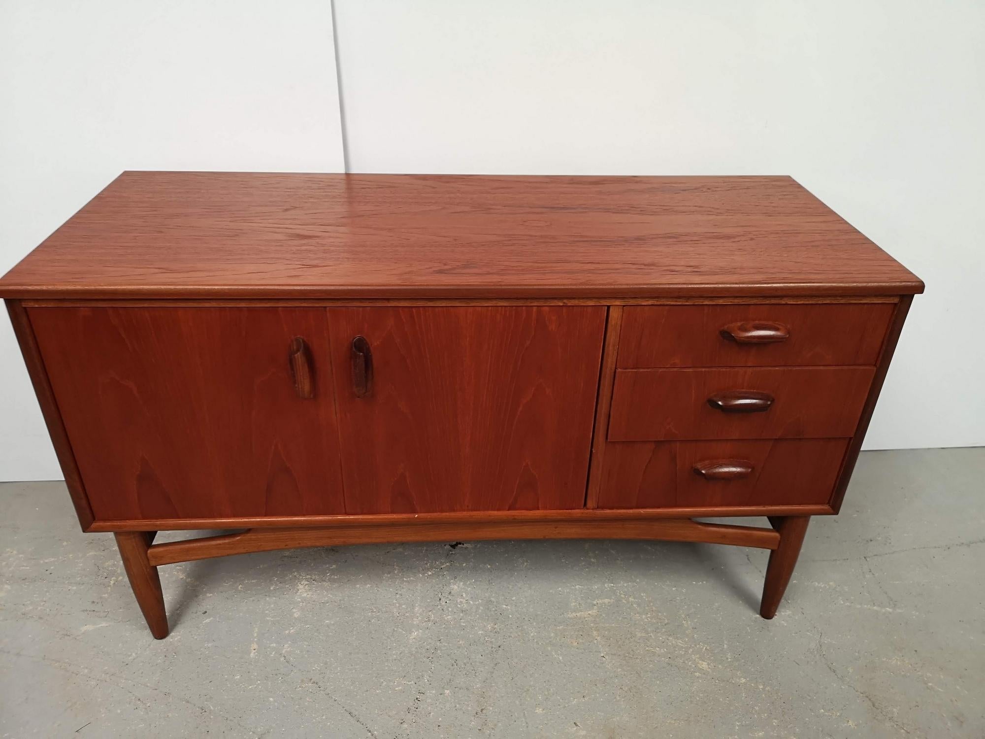 A vintage teak sideboard, its wooden handles are design, quite long.
On the left the doors open onto a space with a shelf.
On the right a series of three drawers of different heights.
The top is rectangular.
The base has a tapered design, with a